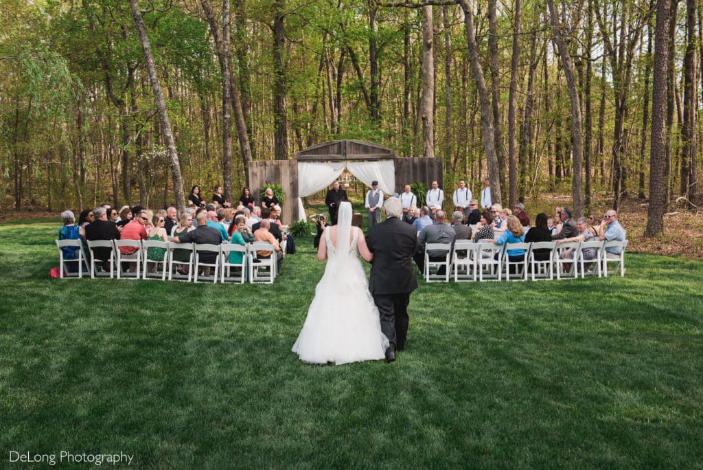 A photograph of the a bride walking down the aisle during an outdoor ceremony at Il Bella Gardens by Charlotte Wedding Photographers DeLong Photography