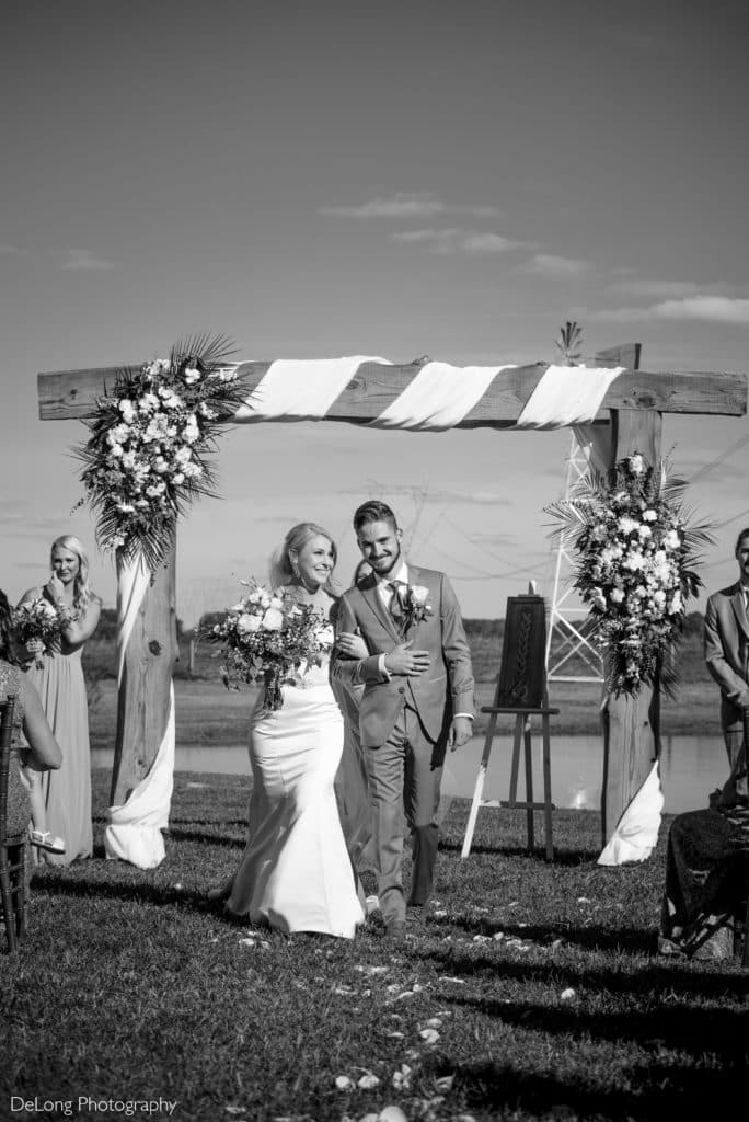 A joyful bride and groom walk arm-in-arm down the aisle after their outdoor wedding ceremony at Dove Meadows. They are framed by a rustic wooden arch adorned with floral arrangements and flowing fabric, set against a serene backdrop of water and open sky. Bridesmaids and guests smile in the background, capturing the celebratory moment. The black-and-white photography by DeLong Photography enhances the timeless and romantic feel of the scene.