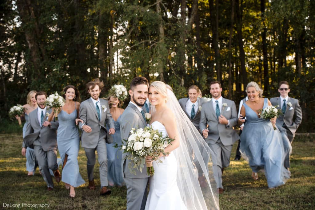 Wedding party running up behind bride and groom for a fun wedding photo at Dove Meadows by Charlotte Wedding Photographers DeLong Photography