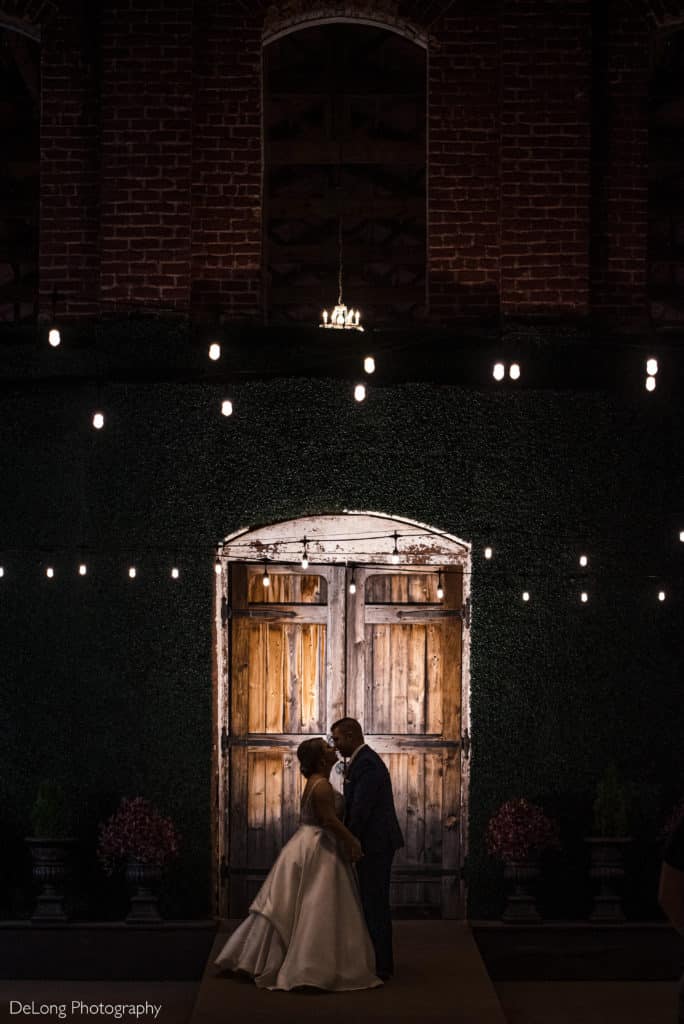 Silhouette of bride and groom about to kiss outside the Providence Cotton Mill doors surrounded by twinkling lights and glassware