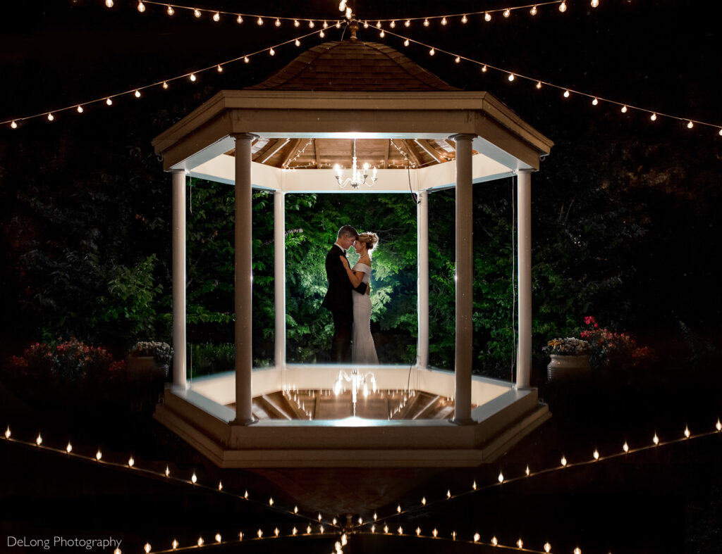 Reflection photograph of bride and groom hugging with their foreheads together in front of greenery withs tring lights above and below from the reflection inside the gazebo on the property of Alexander Homestead in Charlotte, NC by Charlotte wedding photographers DeLong Photography