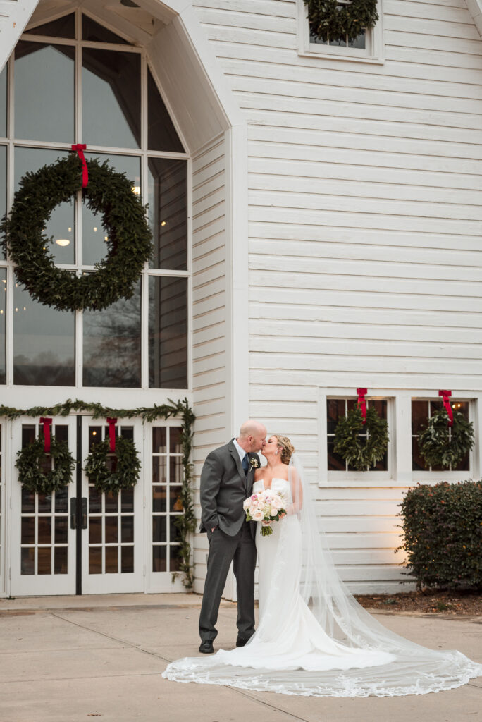 Bride and groom sharing a kiss, the bride's veil and train are beautifully swooped on the ground, outside the Dairy Barn's main entrance that is decorated with large green wreaths and red bows for Christmas time. 