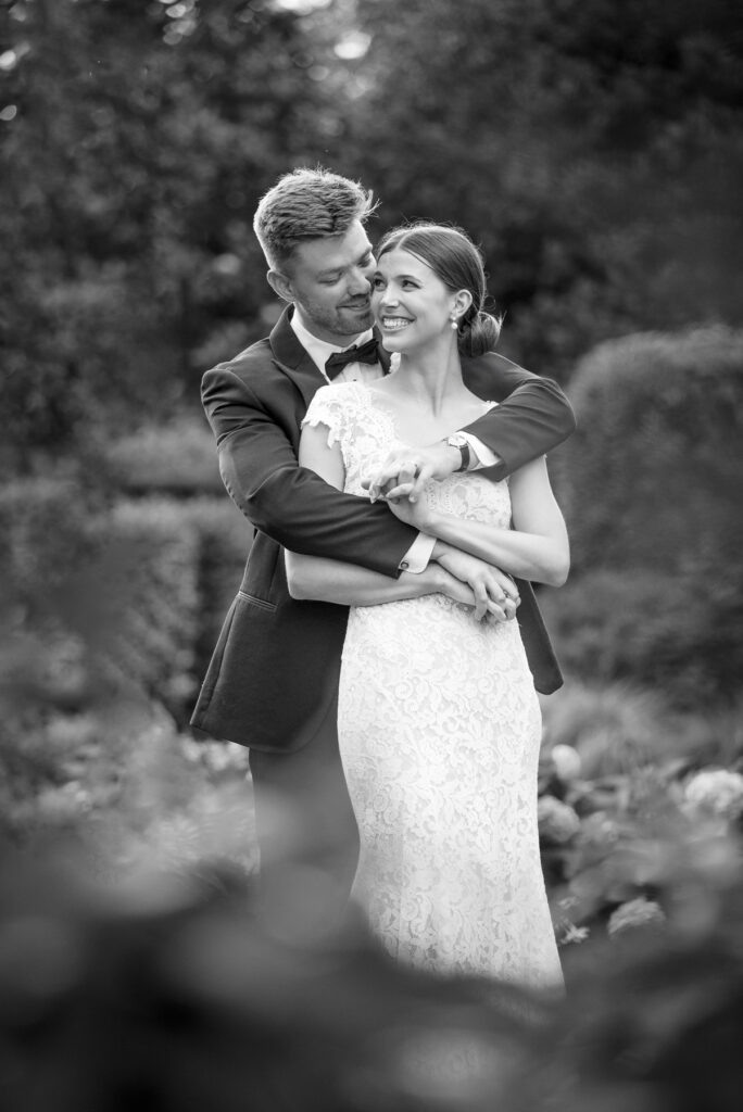 Black and white photograph of a groom hugging his bride from behind. She is looking back toward him giggling. They are standing in a beautifully manicured garden area at Carmel Country Club.