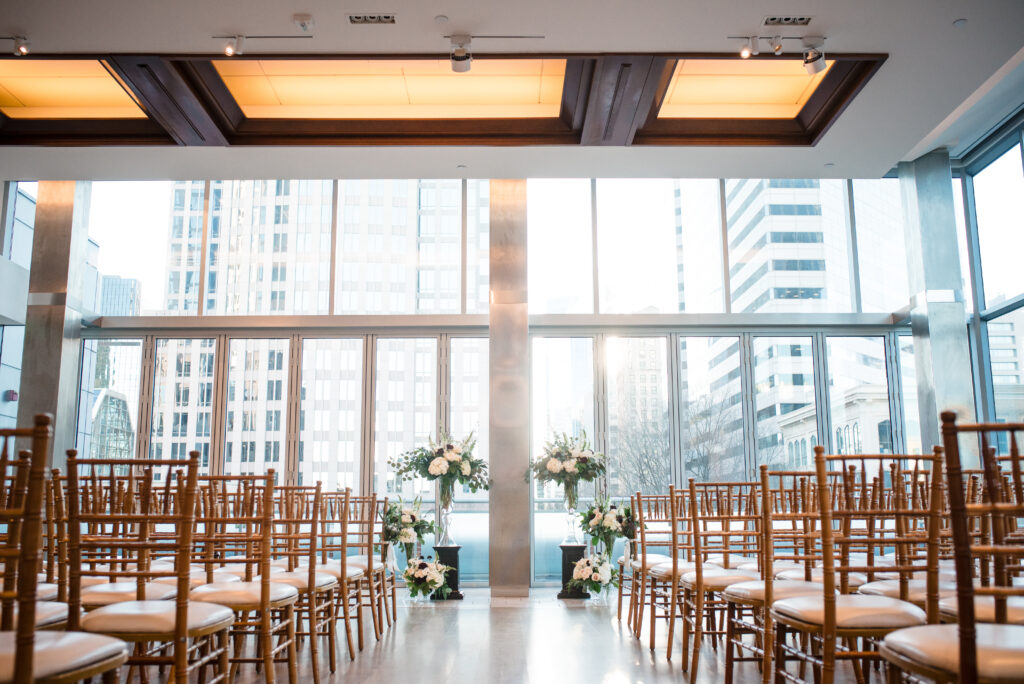 Ceremony setup at Foundation for the Carolinas in Charlotte, NC, featuring rows of wooden Chiavari chairs, elegant floral arrangements, and floor-to-ceiling windows showcasing a stunning city skyline backdrop.