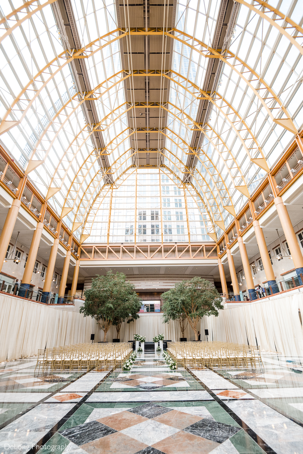 Elegant indoor wedding ceremony setup at Founders Hall in Charlotte, NC, featuring gold chiavari chairs, lush white floral arrangements, and a stunning glass atrium ceiling that floods the venue with natural light. This breathtaking Founders Hall wedding venue is perfect for couples seeking a modern, sophisticated, and unforgettable Charlotte wedding celebration.