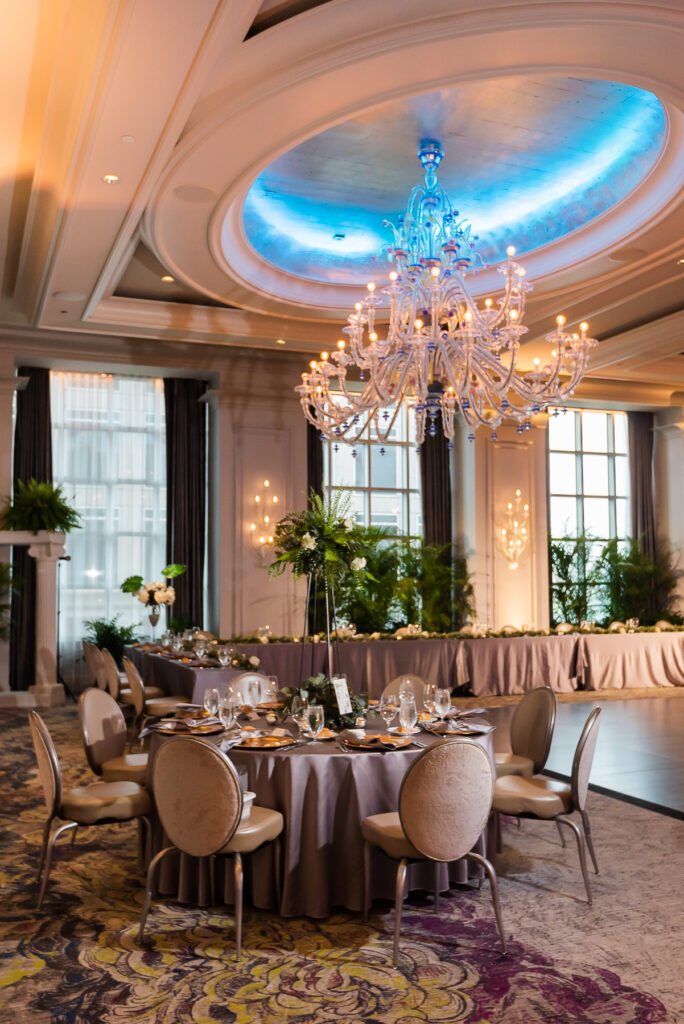 Photograph taken inside the Grand Ballroom at the Grand Bohemian Charlotte. It shows how a couple set up their tables, included lots of greenery in their design, and showcases one of the large chandeliers hanging from the ceiling. 