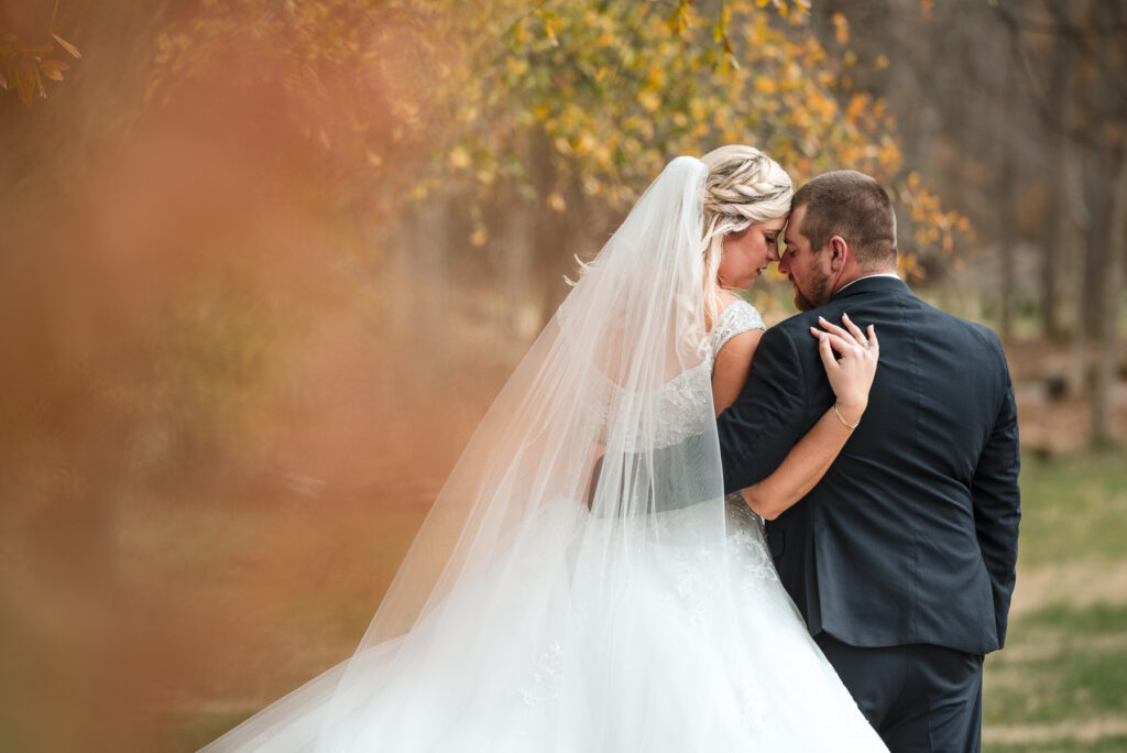 Outdoor portrait of bride and groom facing away from the camera, arms around each other's backs, foreheads together, sharing a peaceful moment amid the yellow and orange foliage of their fall wedding at Low Meadows Estate.