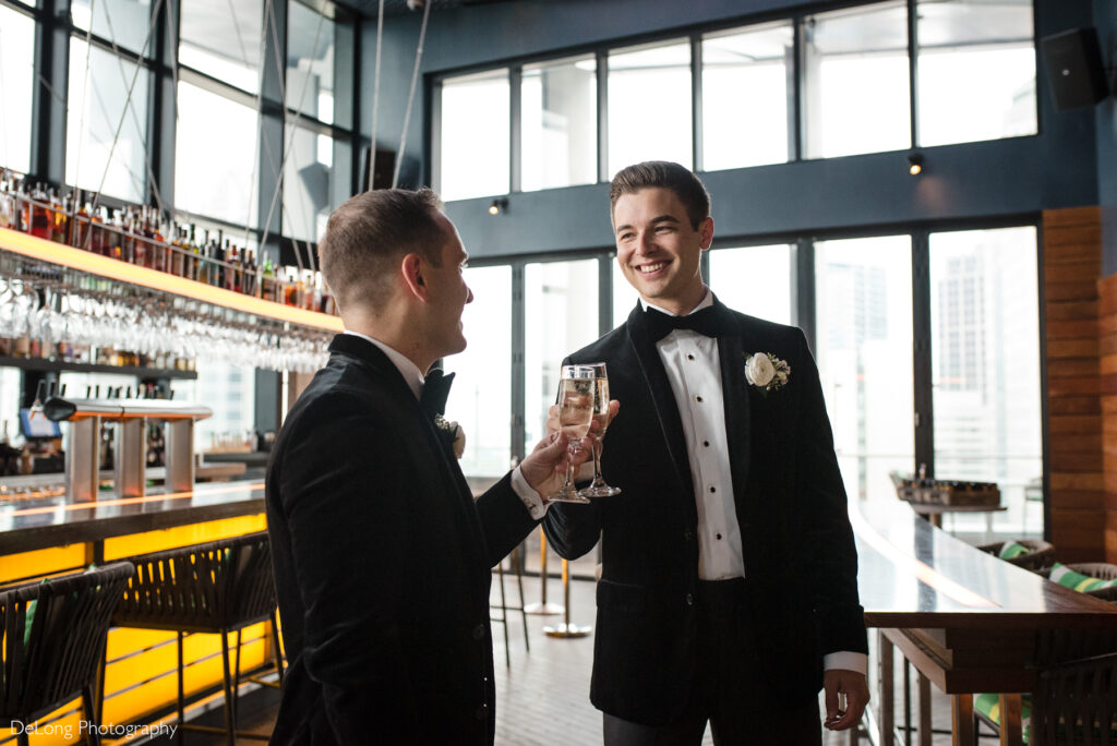 Two grooms in black tuxedos share a champagne toast at Merchant and Trade rooftop bar in Charlotte, NC, before their wedding celebration. This stylish Merchant and Trade wedding venue offers breathtaking skyline views, modern décor, and the perfect backdrop for couples planning a chic Charlotte wedding.  