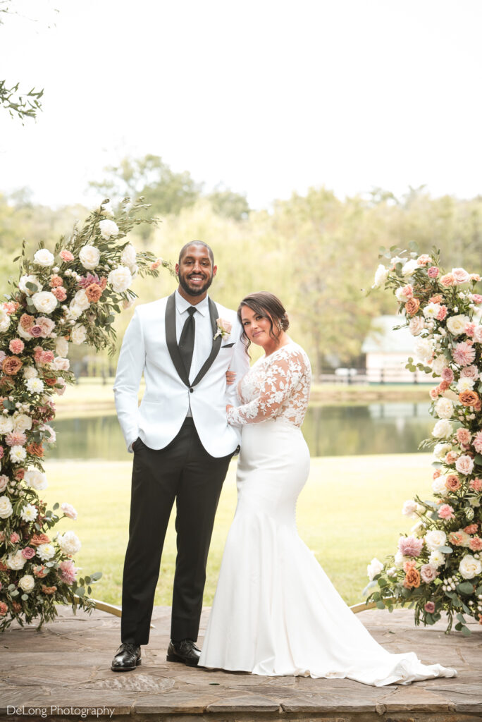 A newlywed couple poses in front of a serene pond at Morning Glory Farms, framed by two lush floral arrangements featuring roses and greenery. The groom, wearing a white tuxedo jacket with black lapels, smiles confidently, while the bride, in a fitted white gown with lace sleeves, leans into him with a soft, joyful expression. The natural backdrop of trees and water enhances the romantic atmosphere. This elegant wedding portrait was captured by DeLong Photography.