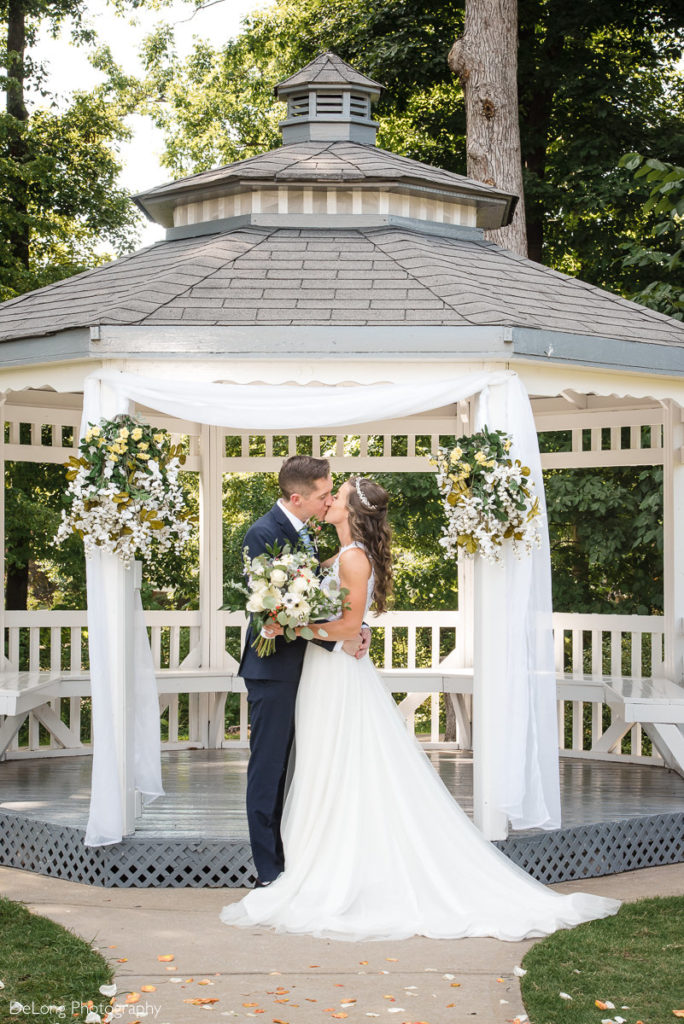 Bride and groom kiss in front of gazebo adorned with flowers at NorthStone Country Club by Charlotte Wedding Photographers DeLong Photography