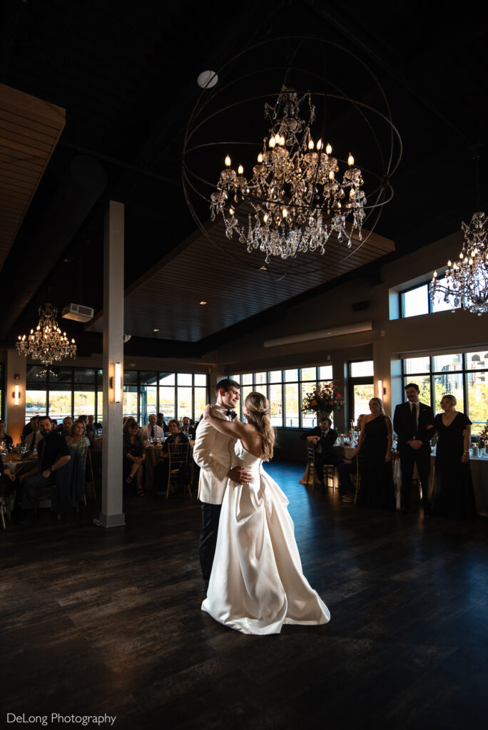 A bride and groom share their first dance at Terrace at Cedar Hill, an elegant wedding venue with floor-to-ceiling windows showcasing a stunning city skyline in the background. The bride wears a classic satin gown with a flowing train, while the groom dons a white tuxedo jacket. The venue features dark wood floors, sparkling crystal chandeliers, and a modern yet romantic ambiance. Wedding guests in formal attire are seated at round tables, watching the couple with warm expressions. Soft natural light filters through the windows, enhancing the intimate and joyous atmosphere. Photo credit: DeLong Photography.