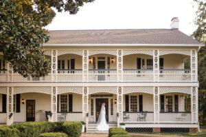 Bridal portrait showcasing the back of the bride's dress as it cascades down the front steps of The White Home. She is standing on the porch looking back over her shoulder at the camera. It is a wide photograph that include the entire front of the home.