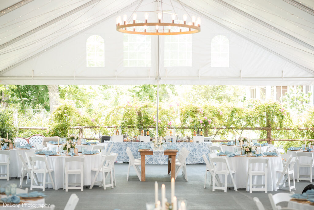 Elegant tented wedding reception setup at The Ivy Place in Lancaster, SC, featuring white chairs, round tables with soft blue accents, lush greenery, and romantic string lights. Captured by Charlotte wedding photographers DeLong Photography.