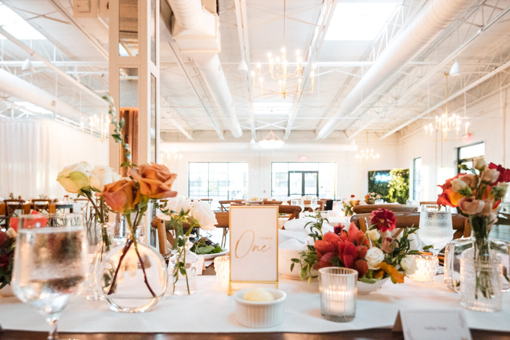 Elegant wedding reception setup at The Ruth by Beau Monde in Charlotte, NC. A beautifully decorated table features delicate floral arrangements in glass vases, glowing candlelight, and a minimalist table number card, with the venue’s airy white interior, large windows, and modern chandeliers in the background.