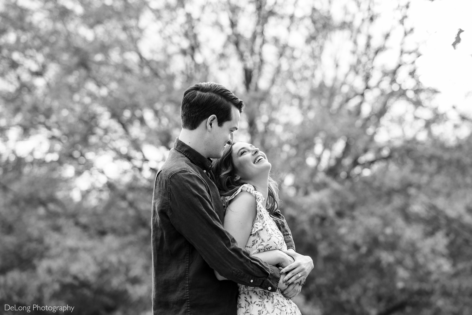 Black and white image of woman looking back at her fiance who is hugging her from behind at a Freedom park in Charlotte, NC by Charlotte Wedding Photographers DeLong Photography