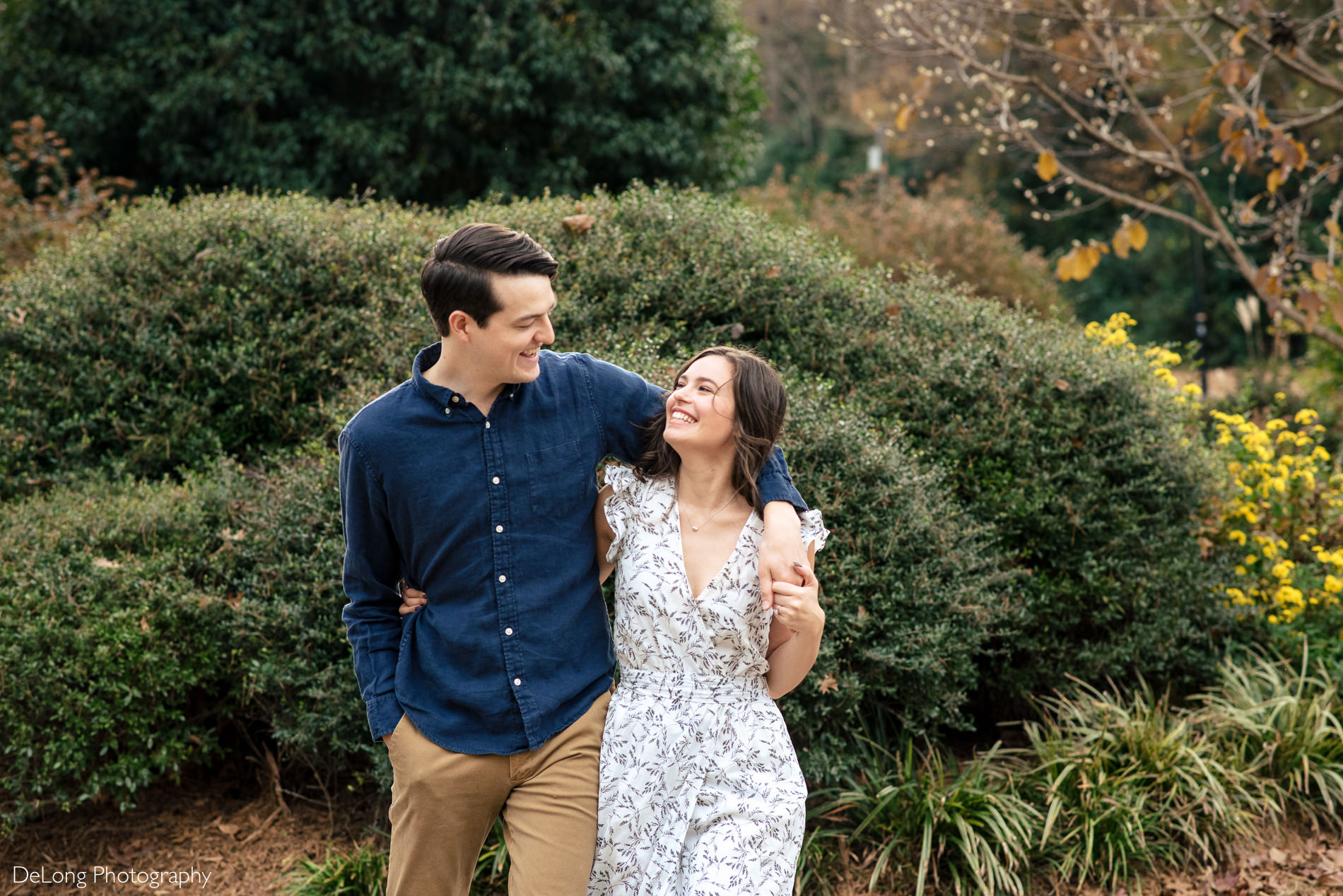 Man with his arm around his fiancee smiling while walking at a Freedom park in Charlotte, NC by Charlotte Wedding Photographers DeLong Photography