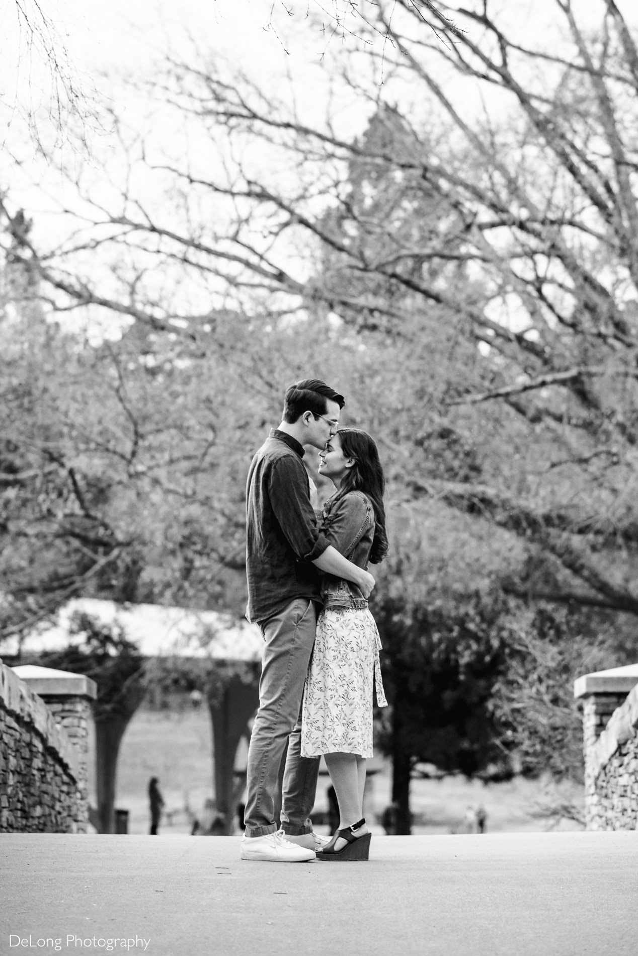Black and white image of man kissing his fiancee on the head standing on the bridge at Freedom Park by Charlotte wedding photographers DeLong Photography