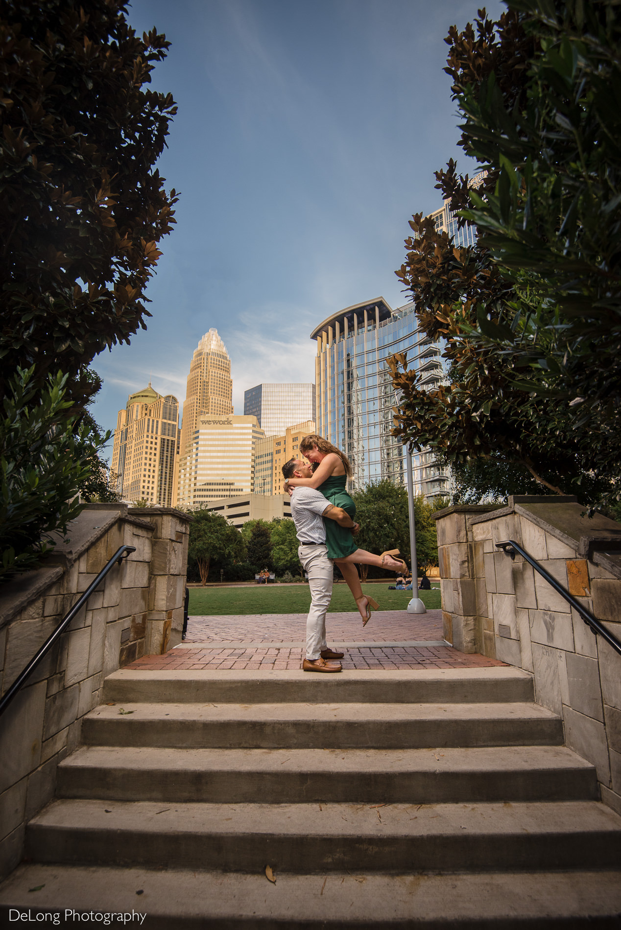 Guy lifting girl up smiling and having fun during a Romare Bearden Park engagement session on a clear blue day with the Charlotte skyline in the background by Charlotte wedding photographers DeLong Photography