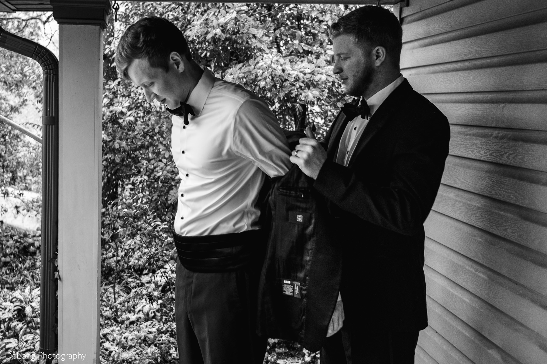 Black and white image of the best man helping the groom out on his jacket on a porch by Charlotte wedding photographers DeLong Photography
