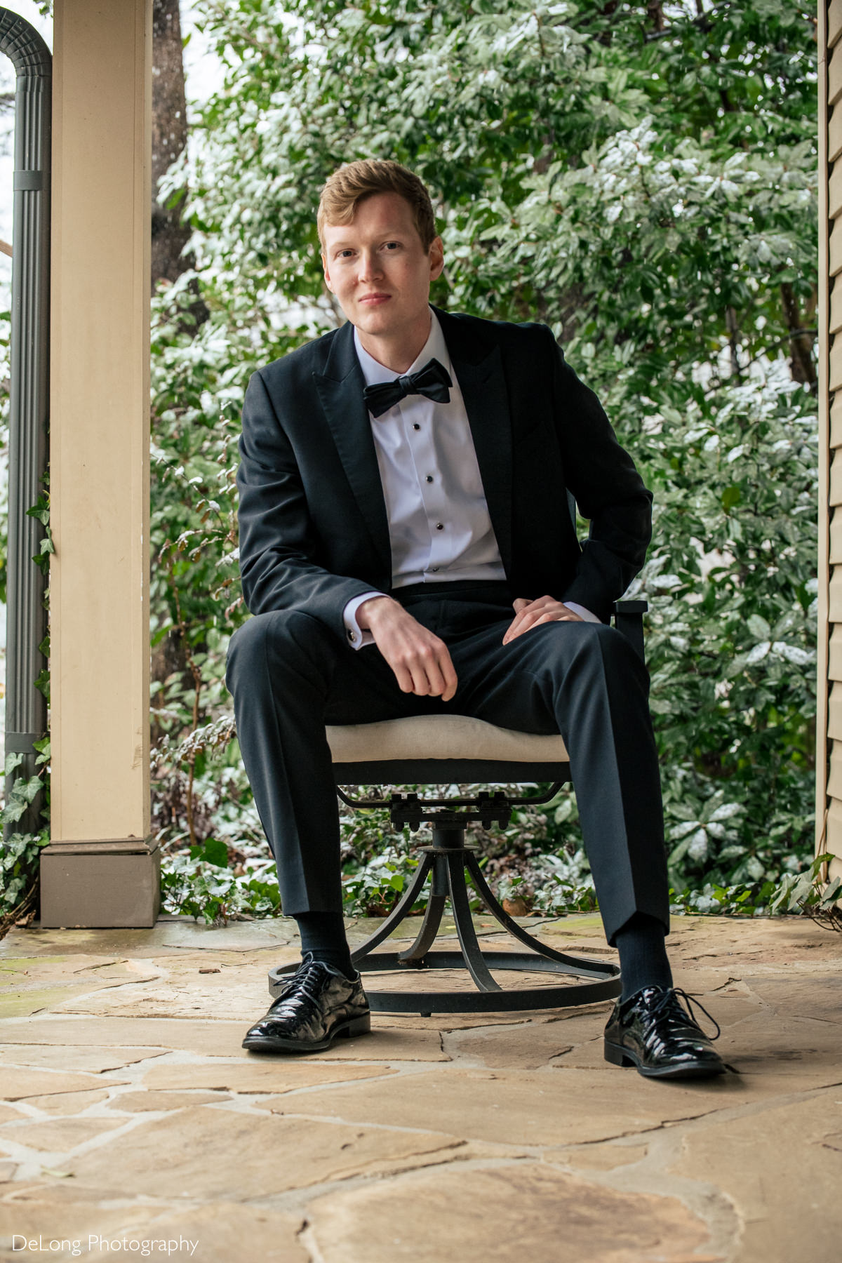 Groom seated portrait on a porch with snow and greenery behind him by Charlotte wedding photographers DeLong Photography