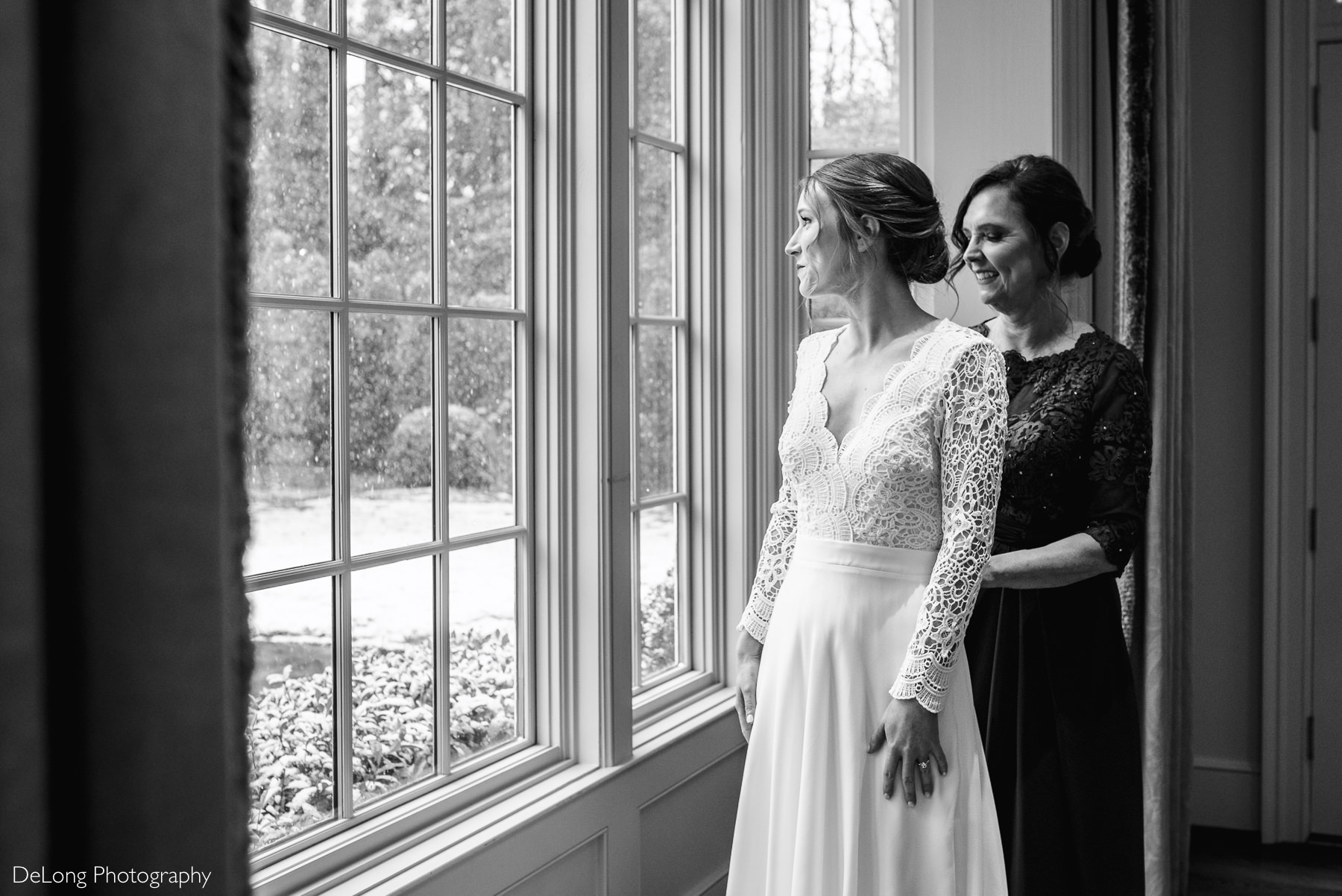 Black and white image of a bride looking out a window as her mother zips up her Lulus wedding dress by Charlotte wedding photographers DeLong Photography