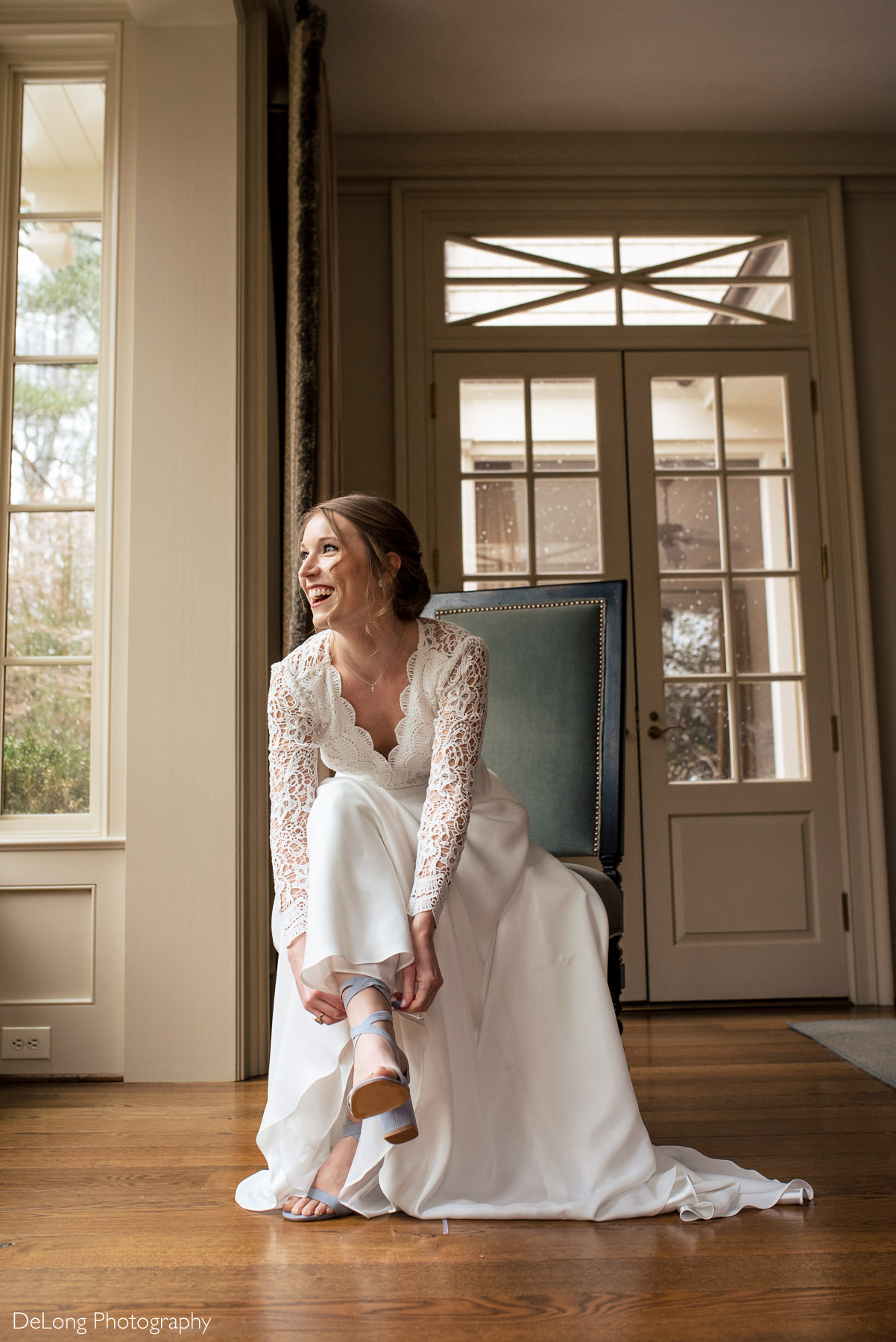 Seated portrait of a bride smiling and laughing while tying her cornflower blue wedding shoes by Charlotte wedding photographers DeLong Photography