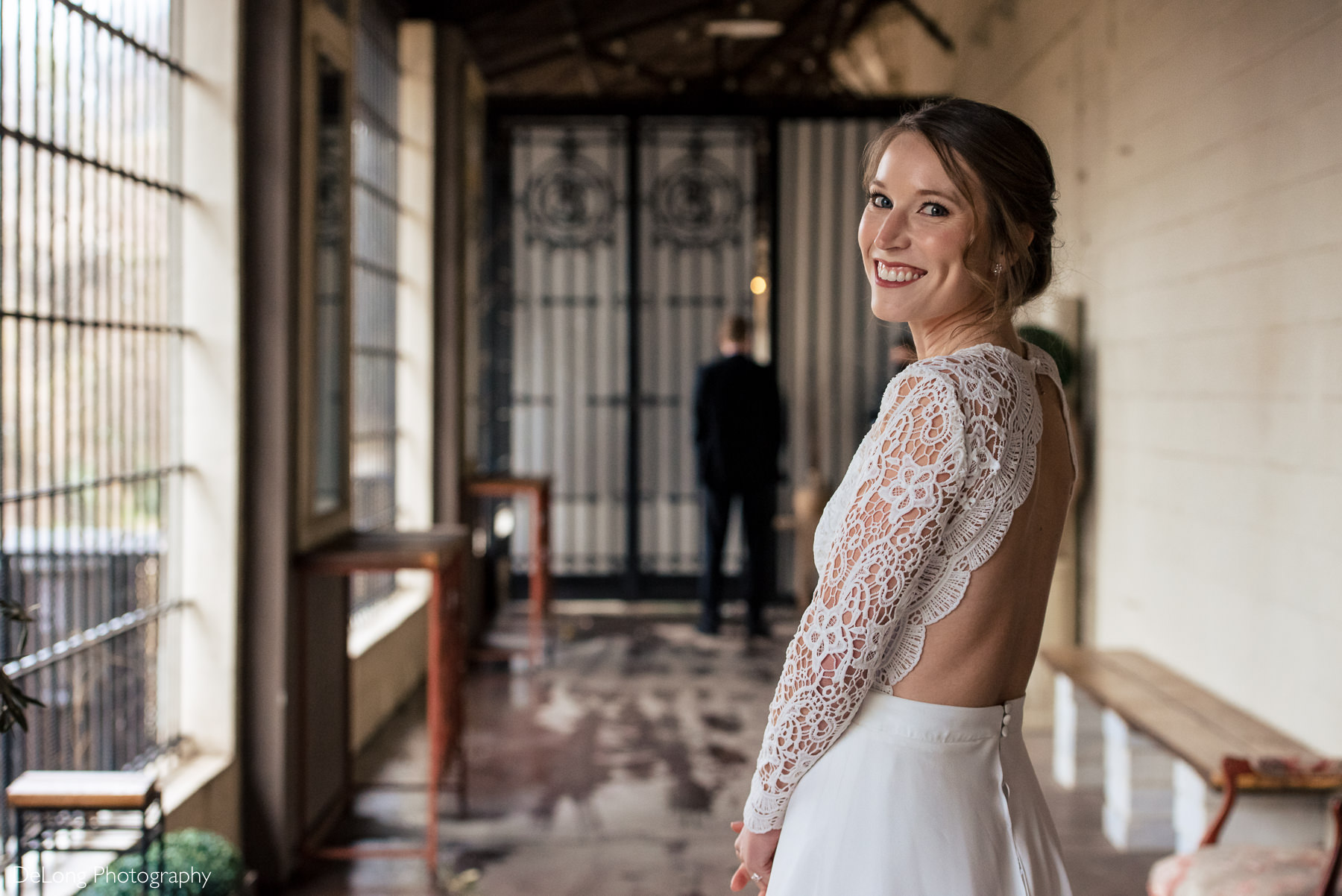 A groom blurred in the background as a bride smiles looking back at the camera before a first look by Charlotte wedding photographers DeLong Photography
