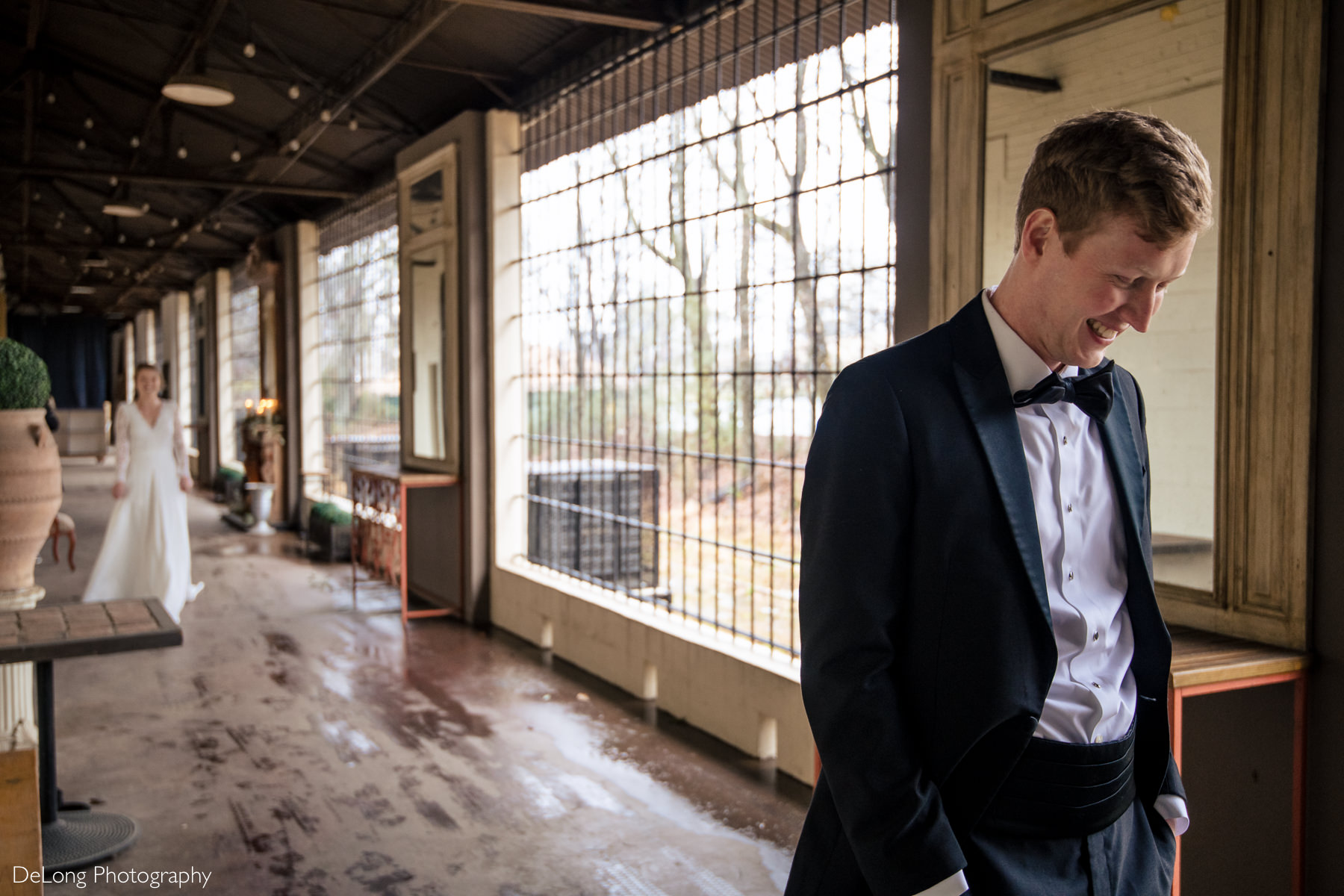 A bride blurred in the background walking up behind a smiling groom before a first look at the Westside Warehouse in Atlanta by Charlotte wedding photographers DeLong Photography