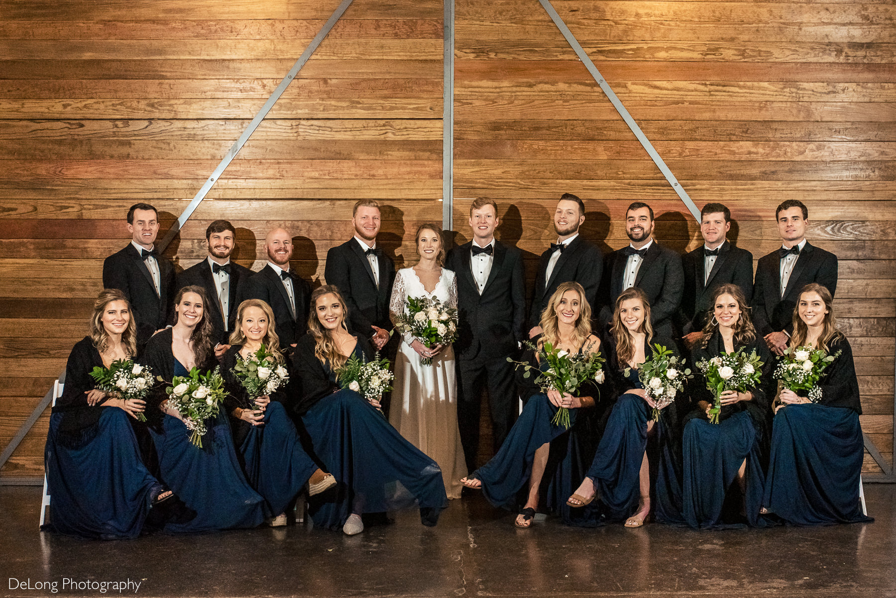 Full wedding party portrait where the bridesmaids are seated and the groomsmen are standing behind them in front of the wooden doors at the Westside warehouse in Atlanta by Charlotte wedding photographers DeLong Photography