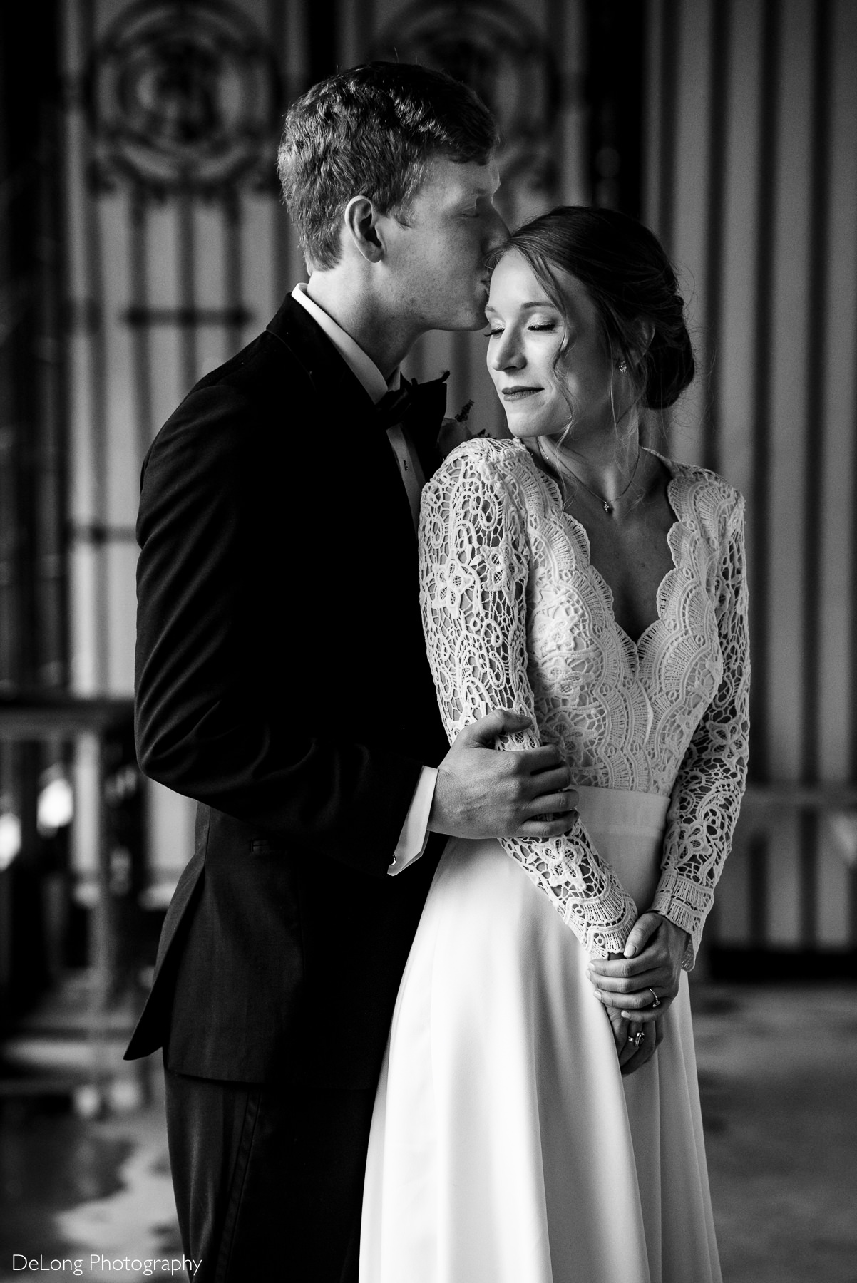 Black and white photograph of a bride softly smiling standing in front of her groom looking back toward him with her eyes closed as he kisses her temple sweetly by Charlotte wedding photographers DeLong Photography