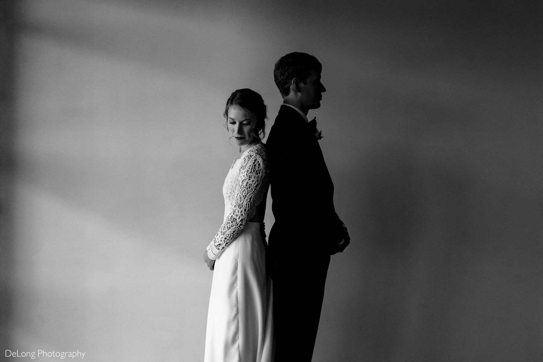 Bride and groom positioned in front of a smooth blank wall back to back the groom silhouetted and the bride Rembrandt lit as she looks back toward the groom by Charlotte wedding photographers DeLong Photography
