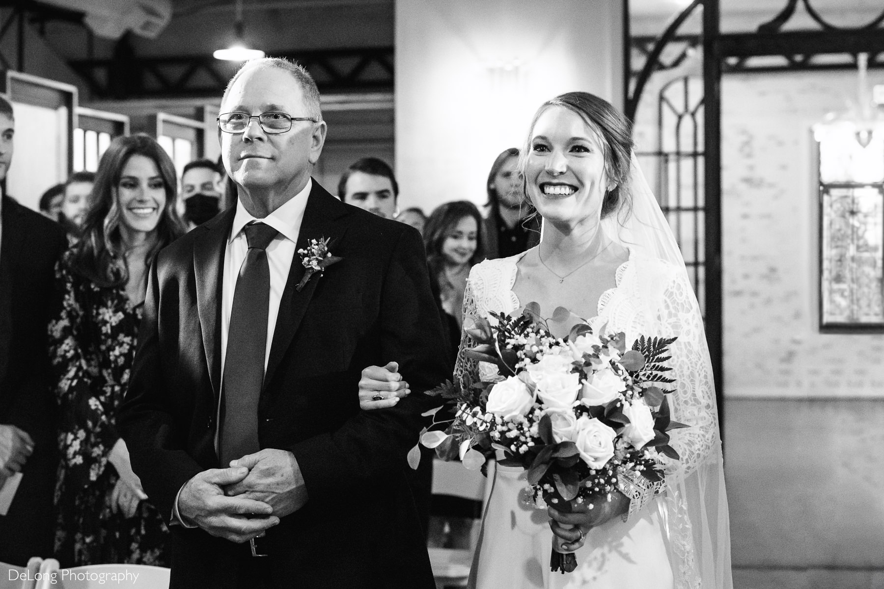 Black and white image of a bride beaming as she comes down the aisle with her father at the Westside Warehouse by Charlotte wedding photographers DeLong Photography