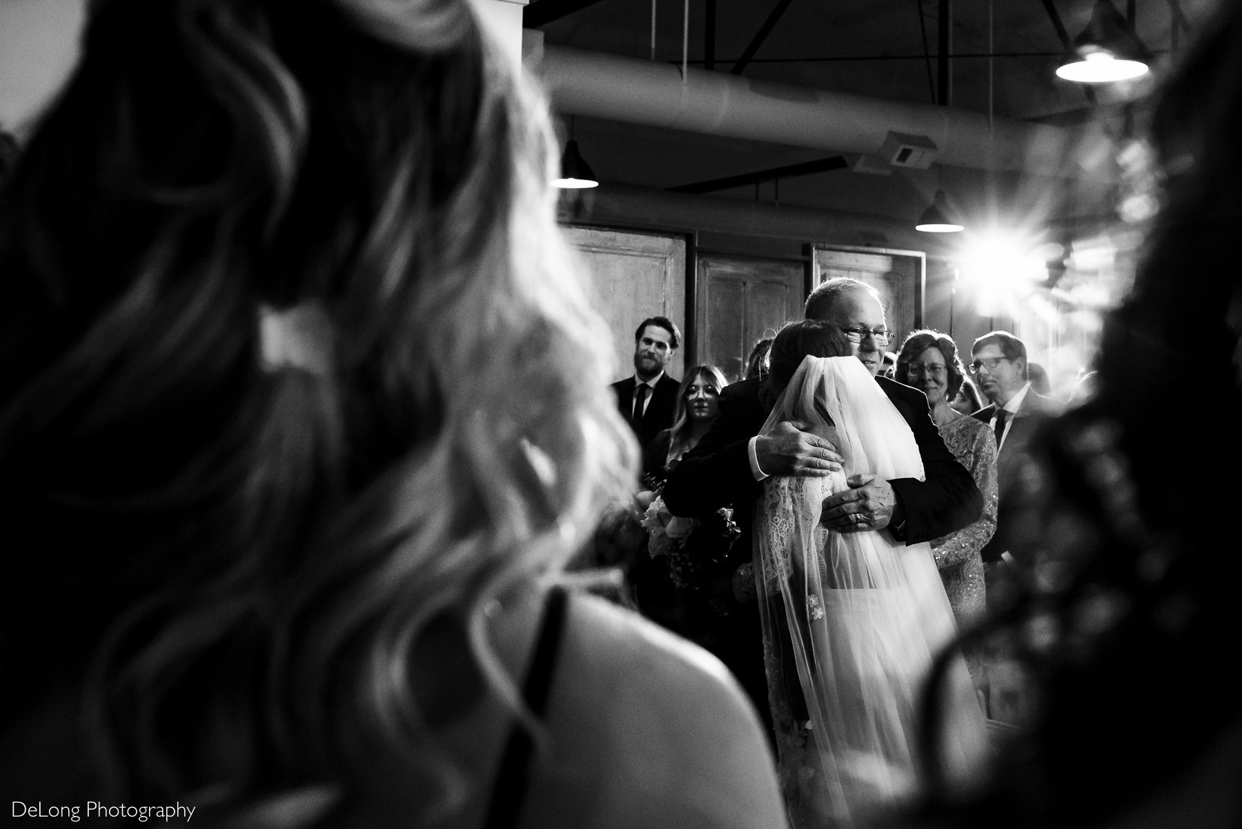 Photograph taken between two bridesmaids at the front of a wedding ceremony of a father and bride hugging as he gives her away by Charlotte wedding photographers DeLong Photography