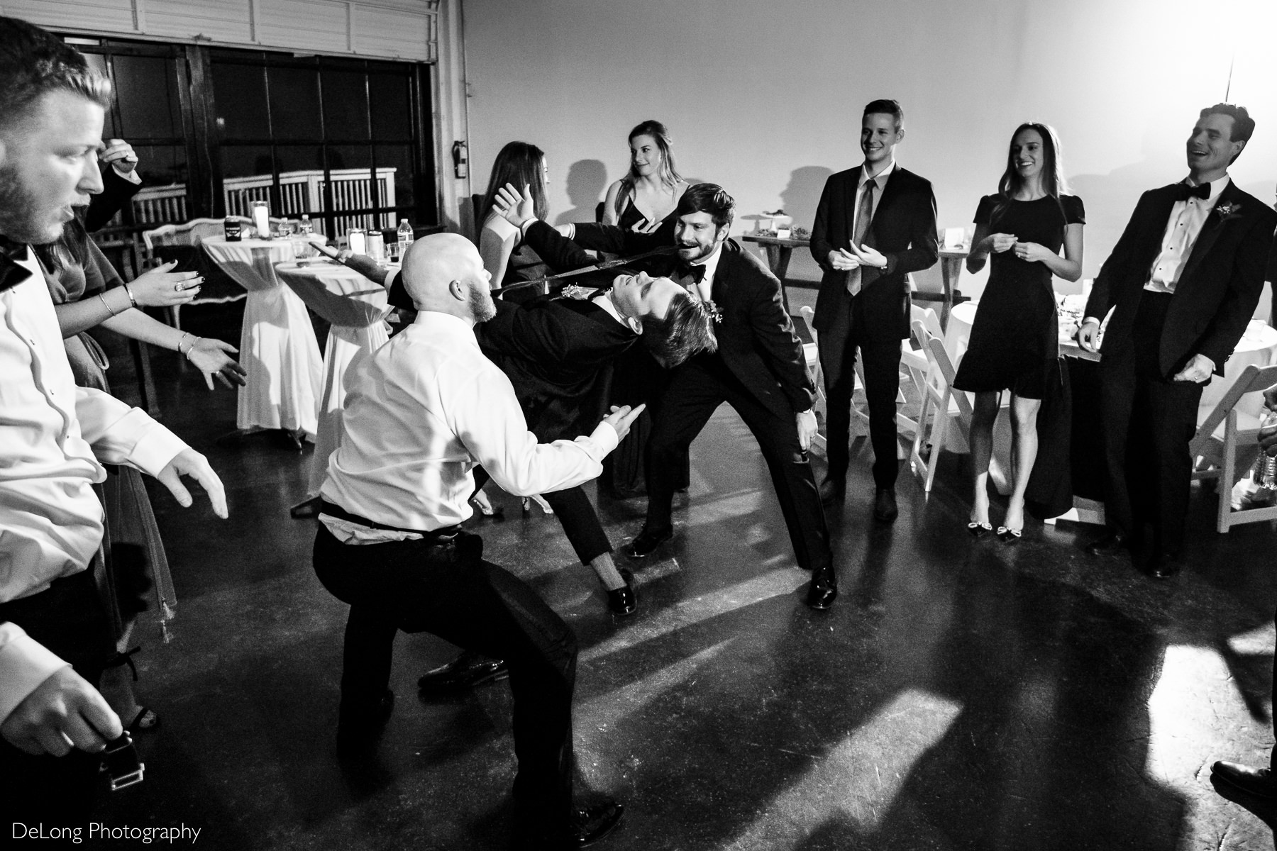 Black and white image of a man bent backwards mid walk doing the limbo under a tie that is being held in the mouths of a groomsmen on each end at the Westside warehouse in Atlanta by Charlotte wedding photographers DeLong Photography
