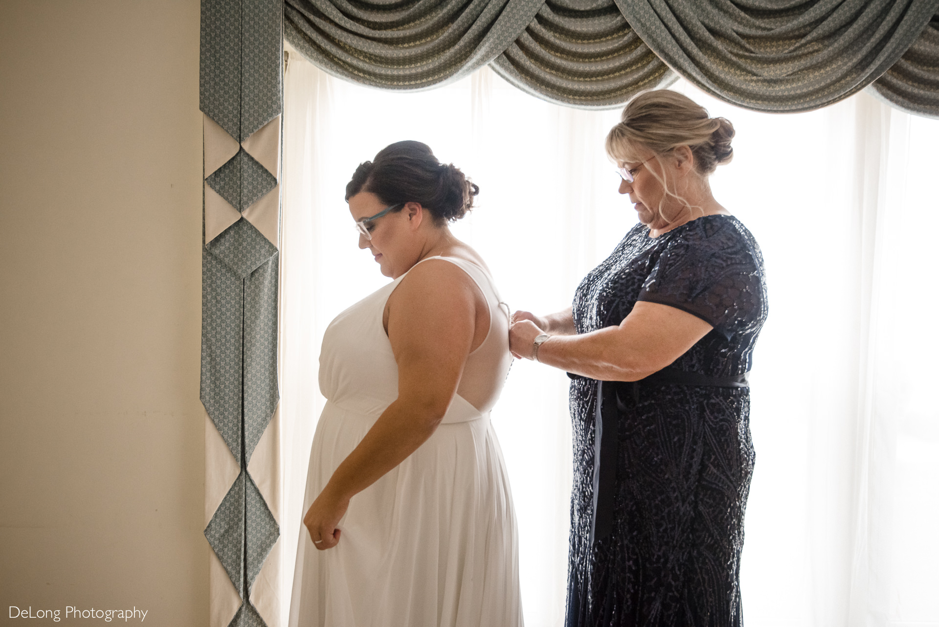 Bride having her dress buttoned up by her mother near a window at the Island House by Charlotte Wedding Photographers DeLong Photography