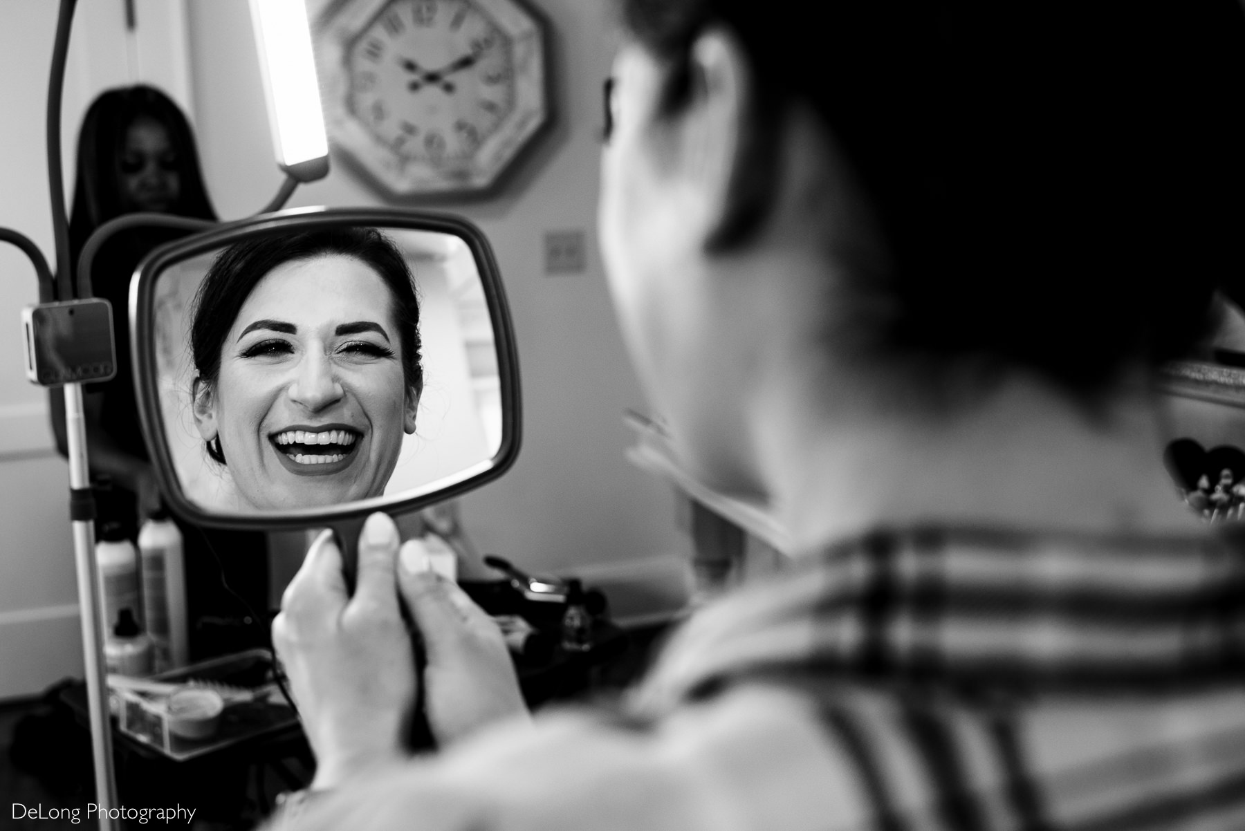Black and white image of a reflection showing bride smiling while looking into make up mirror by Charlotte Wedding Photographers DeLong Photography
