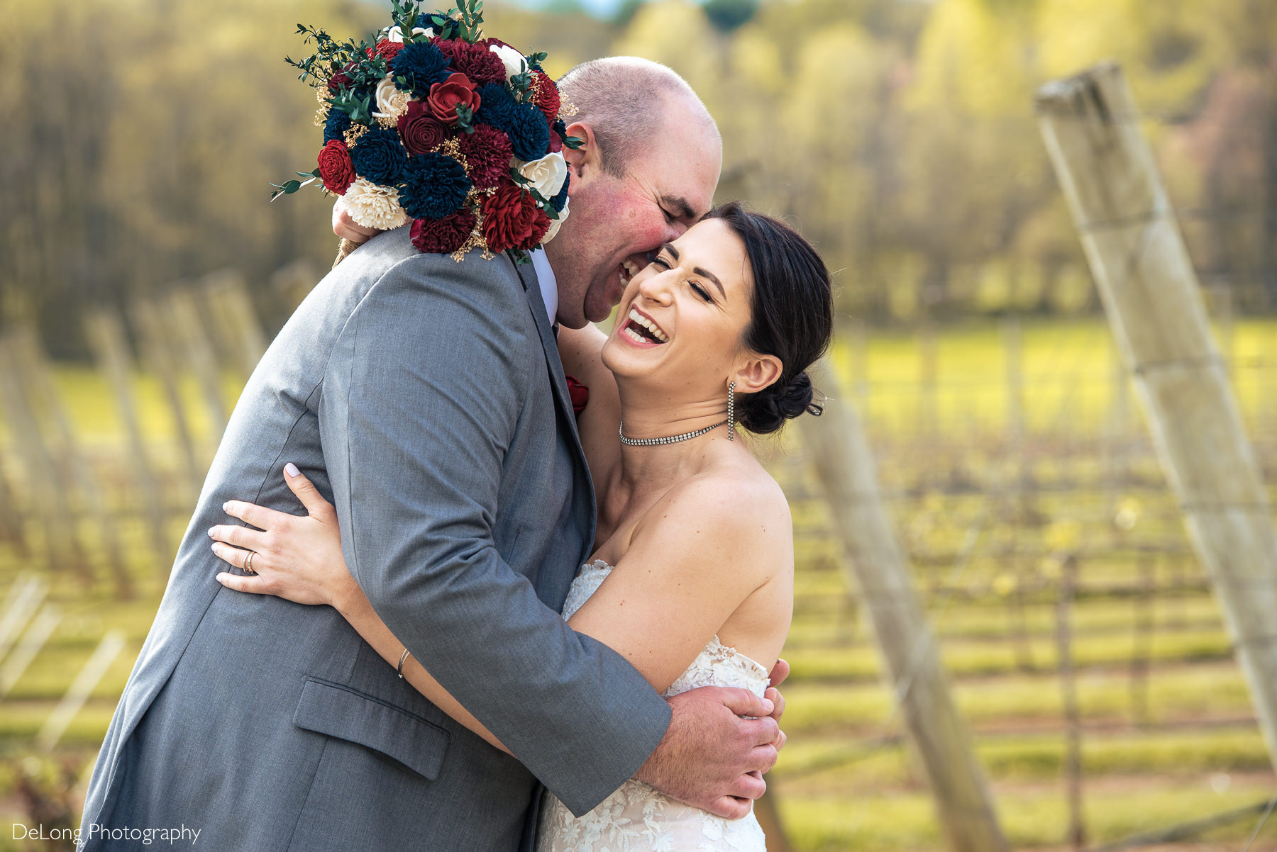 Bride and groom laughing with red, white, and navy bouquet over groom's shoulder during portraits at Childress Vineyards by Charlotte Wedding Photographers DeLong Photography