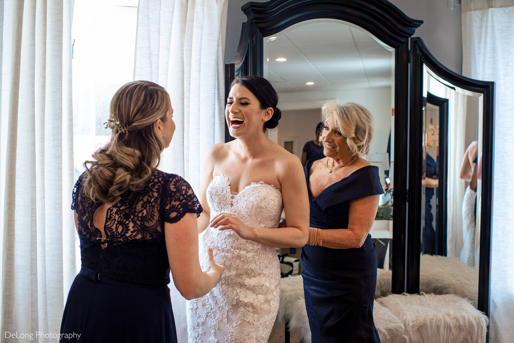 Bride laughing with Maid of Honor while the Mother of the Bride zips up her dress in the bridal suite at Childress Vineyards by Charlotte Wedding Photographers DeLong Photography