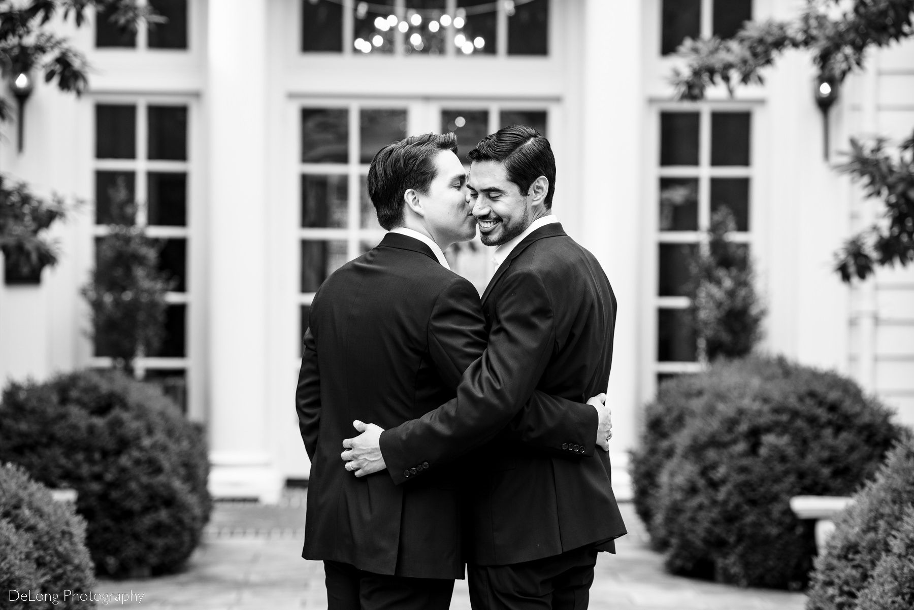 Black and white portrait of a groom smiling while getting a kiss on his cheek from his husband at the Duke Mansion by Charlotte wedding photographers DeLong Photography