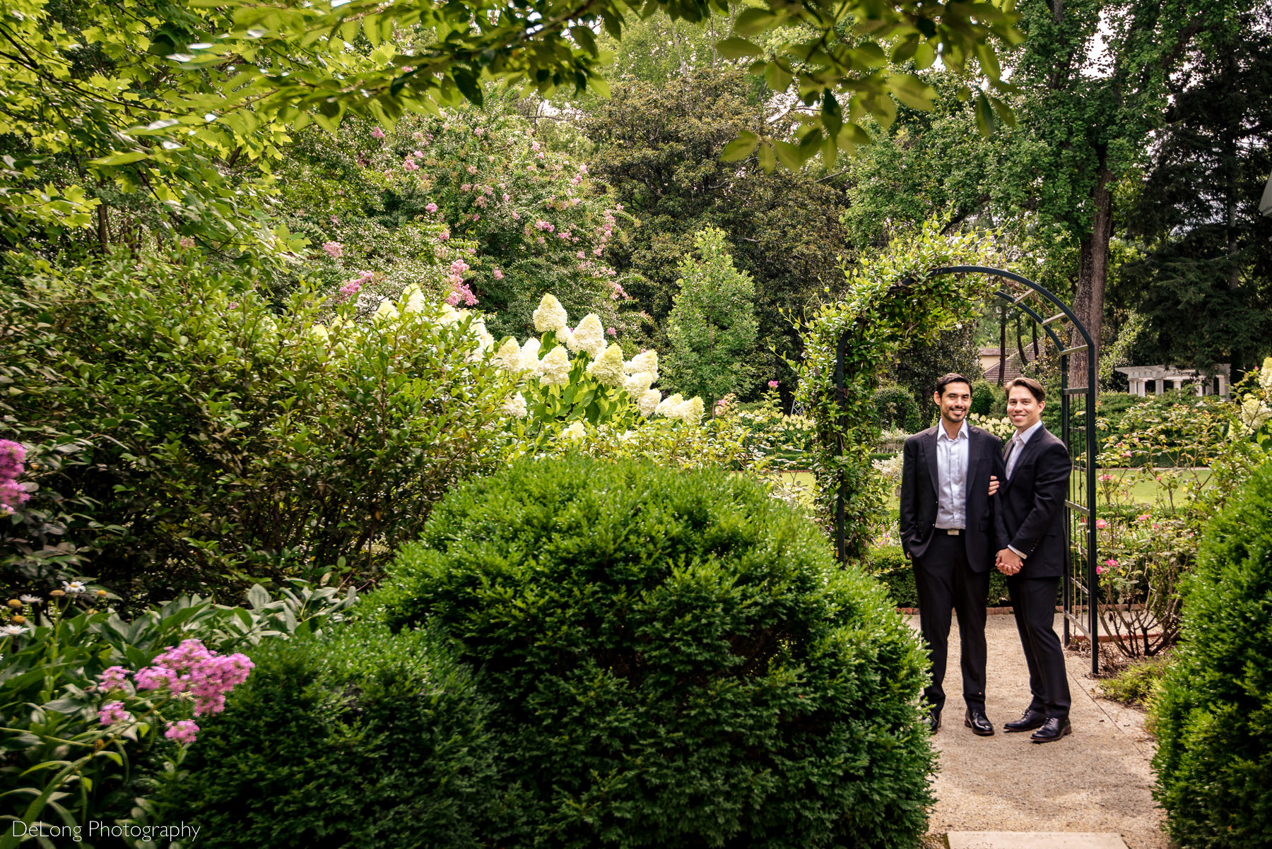Two grooms smiling at the camera being framed by greenery and flowers at the Duke Mansion by Charlotte wedding photographers DeLong Photography