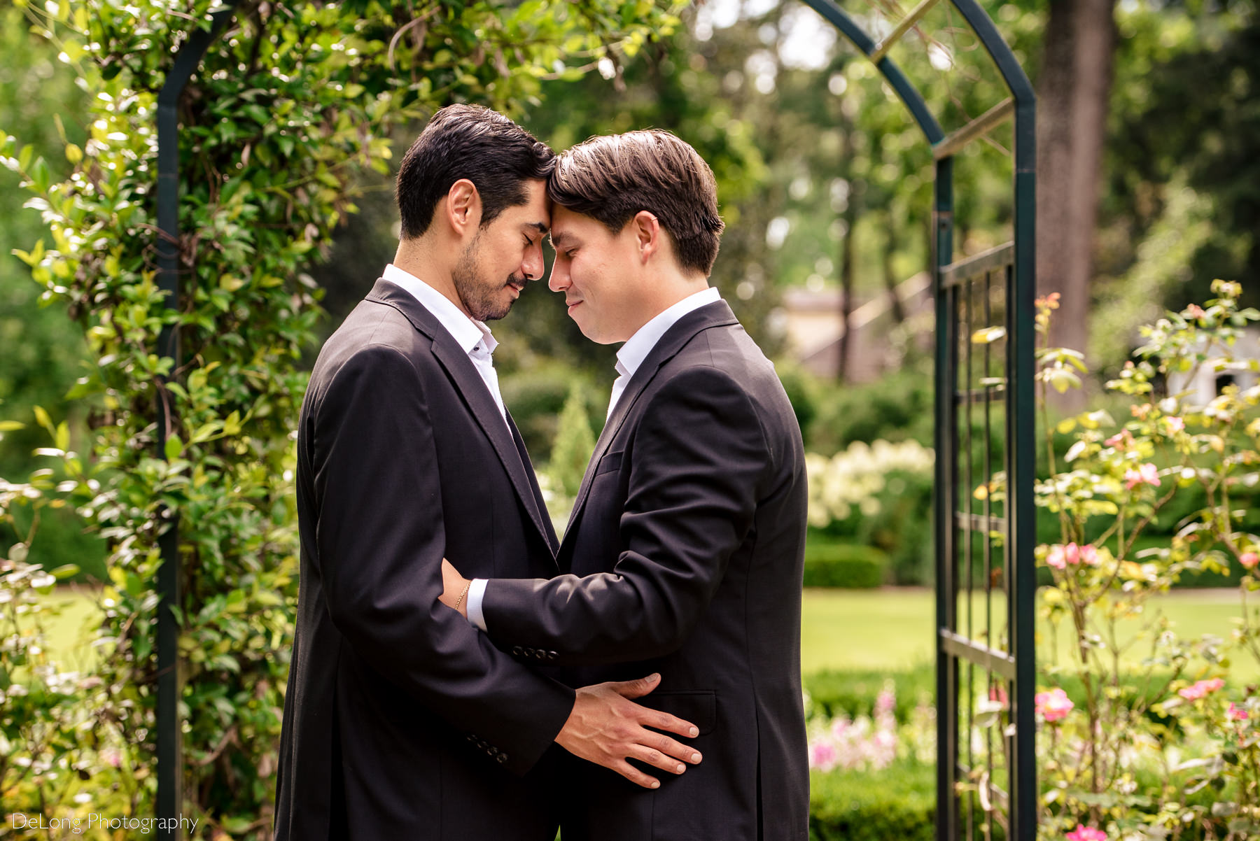 Same-sex couple holding one another with their foreheads together underneath a garden arbor at the Duke Mansion by Charlotte wedding photographers DeLong Photography