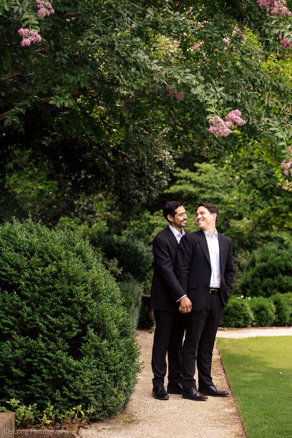 Two grooms holding hands looking at one another framed by flowers and greenery on the day of their summer elopement at the Duke Mansion by Charlotte wedding photographers DeLong Photography
