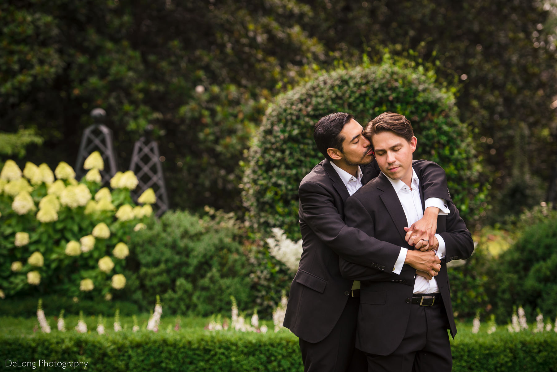 Serene and sweet portrait of a groom getting a kiss on the cheek from his husband in the gardens of the Duke Mansion by Charlotte wedding photographers DeLong Photography