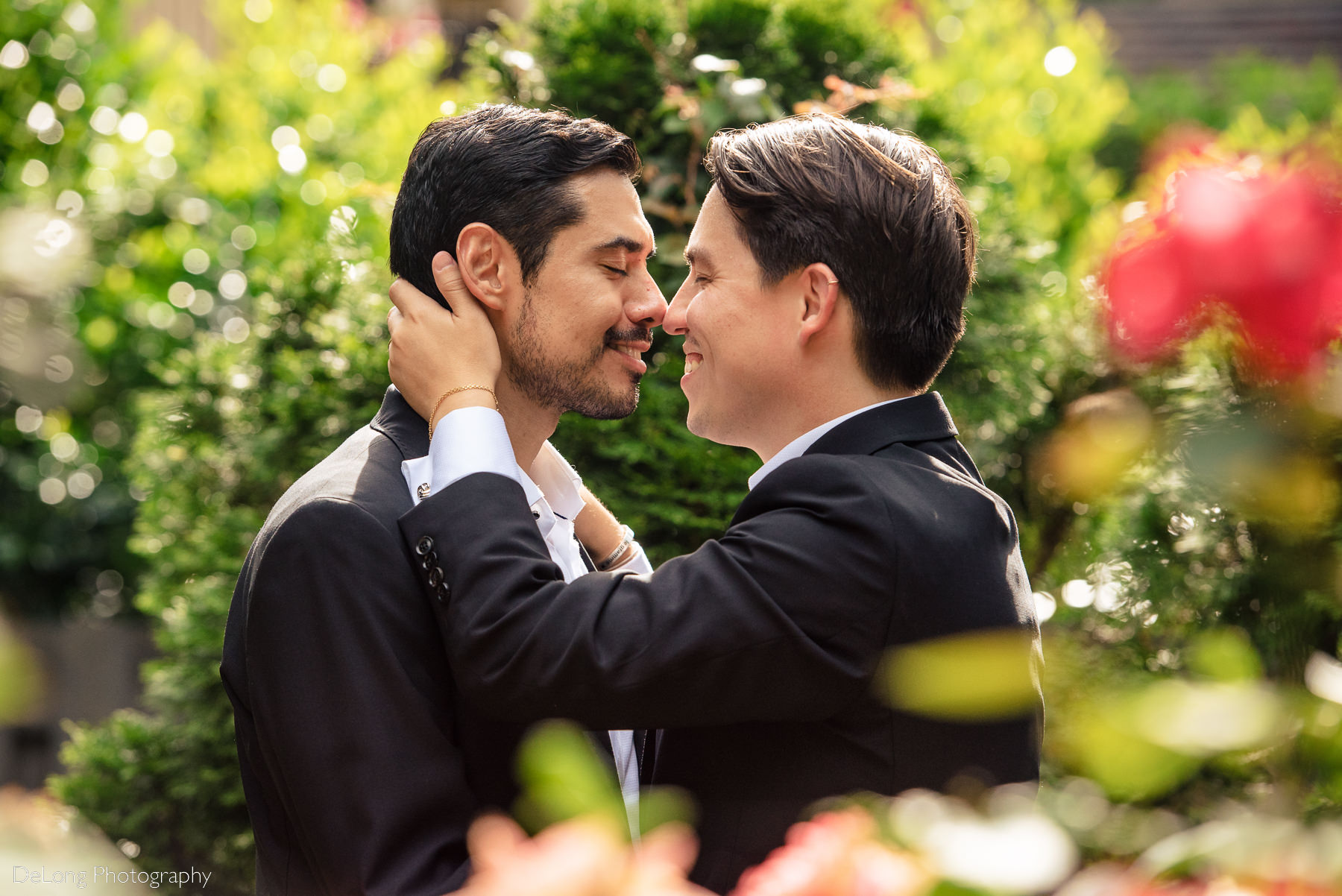 Portrait with bokeh leaves and flowers in the foreground of two grooms gently giving eskimo kisses in the gardens of the Duke Mansion by Charlotte wedding photographers DeLong Photography