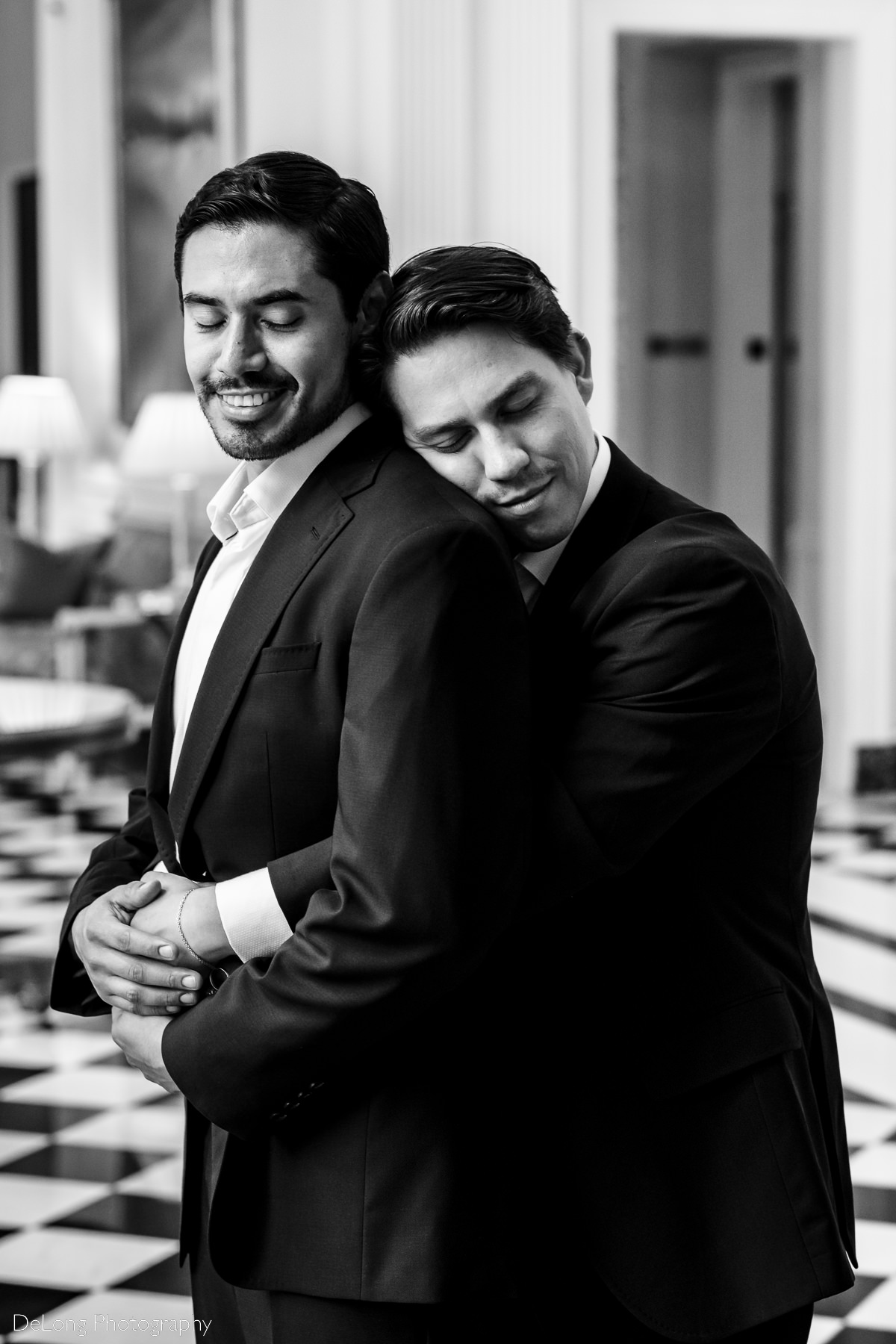 Black and white portrait of a groom smiling while receiving a hug from his husband from behind inside the Duke Mansion by Charlotte wedding photographers DeLong Photography