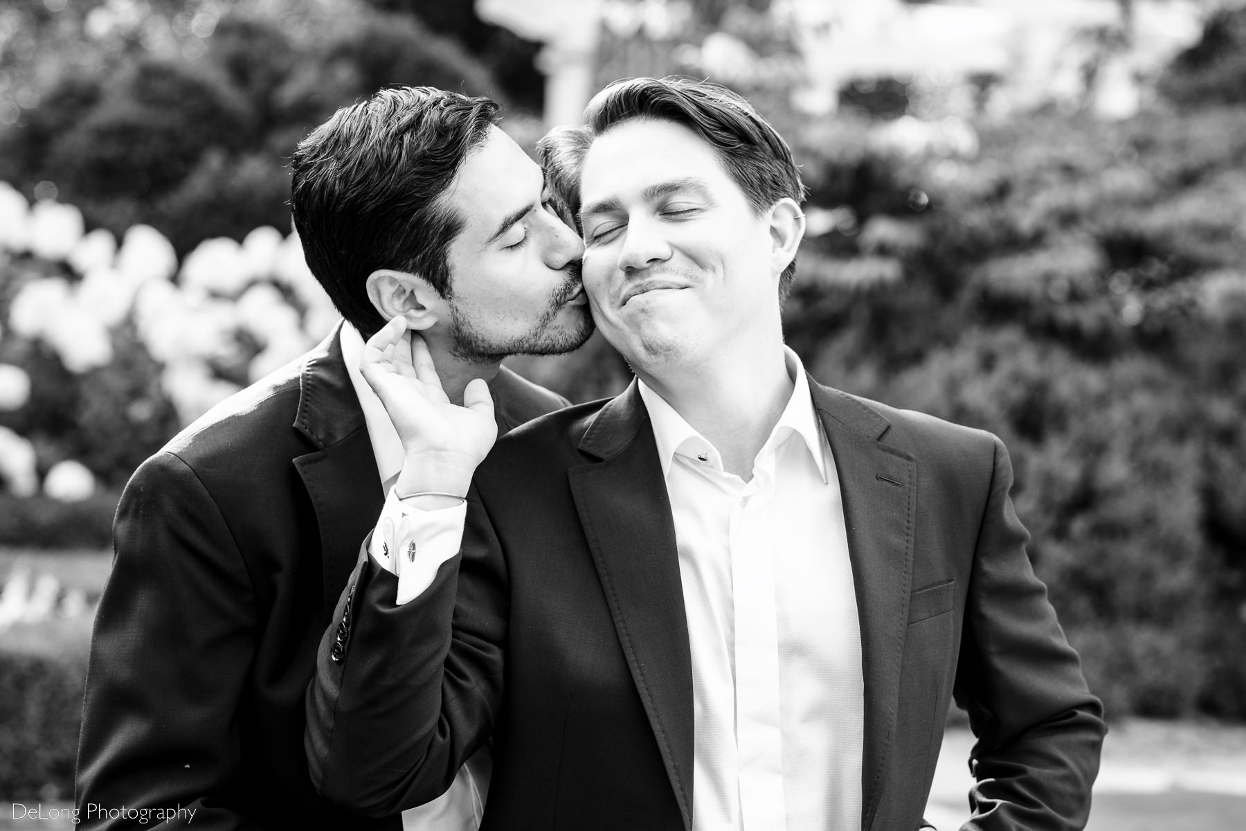 Black and white portrait of groom smiling with his eyes closed touching the face of his husband who is kissing him from behind by Charlotte wedding photographers DeLong Photography