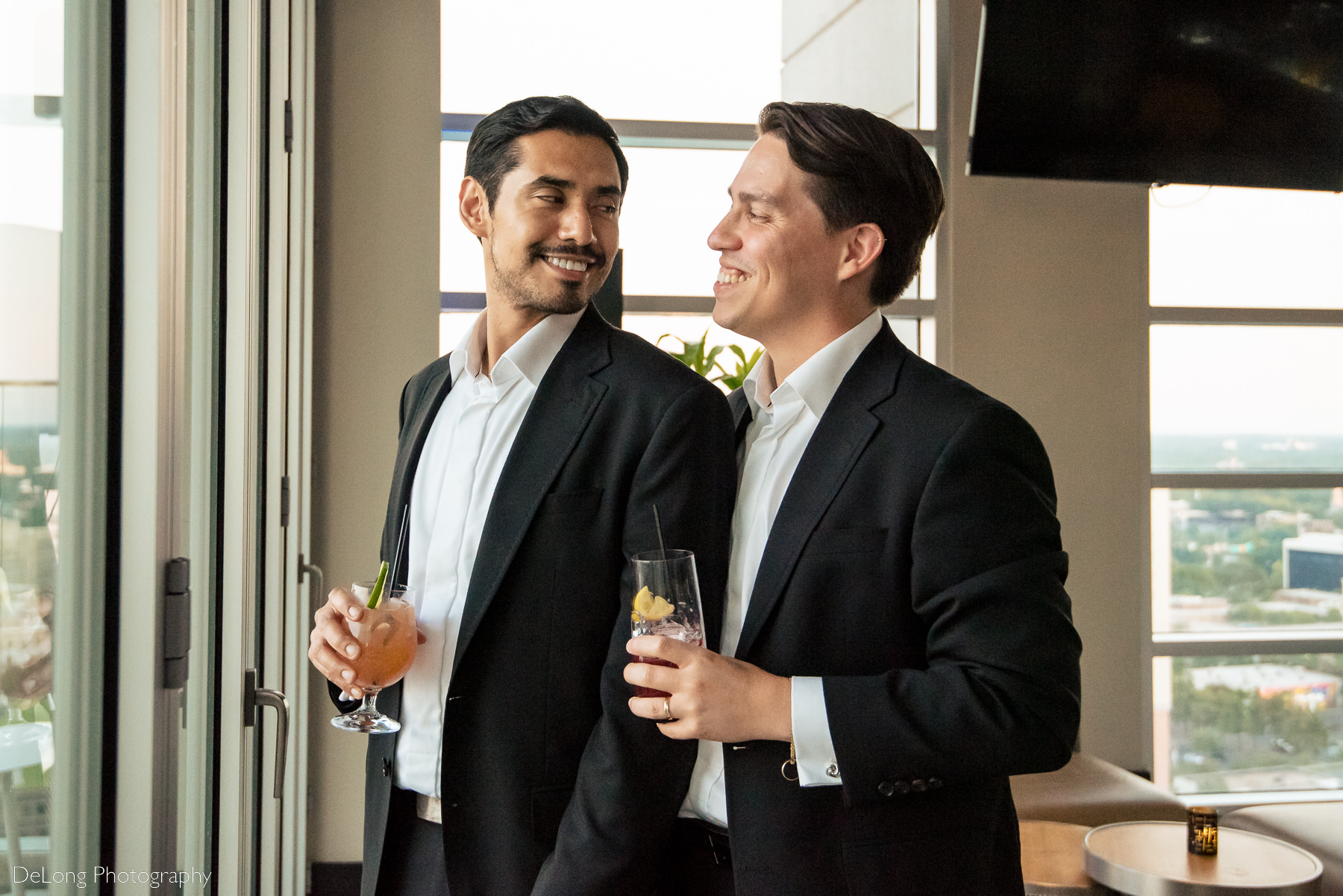 Two grooms smiling at one another holding cocktails inside Nuvole 22 by Charlotte wedding photographers DeLong Photography