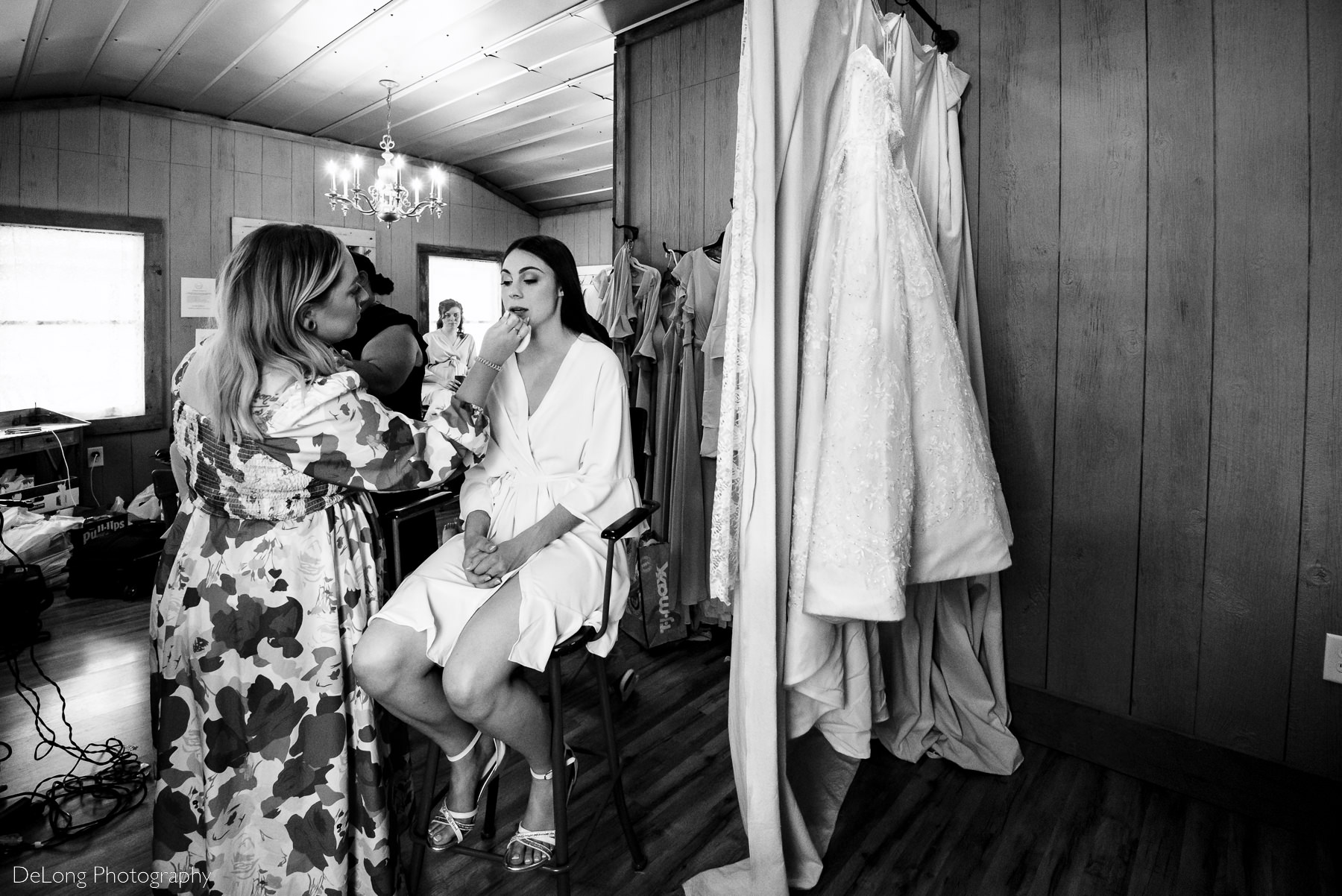 Black and white photograph of a bride getting her makeup done while simultaneously showing her wedding dress hidden behind a curtain by Charlotte Wedding Photographers DeLong Photography