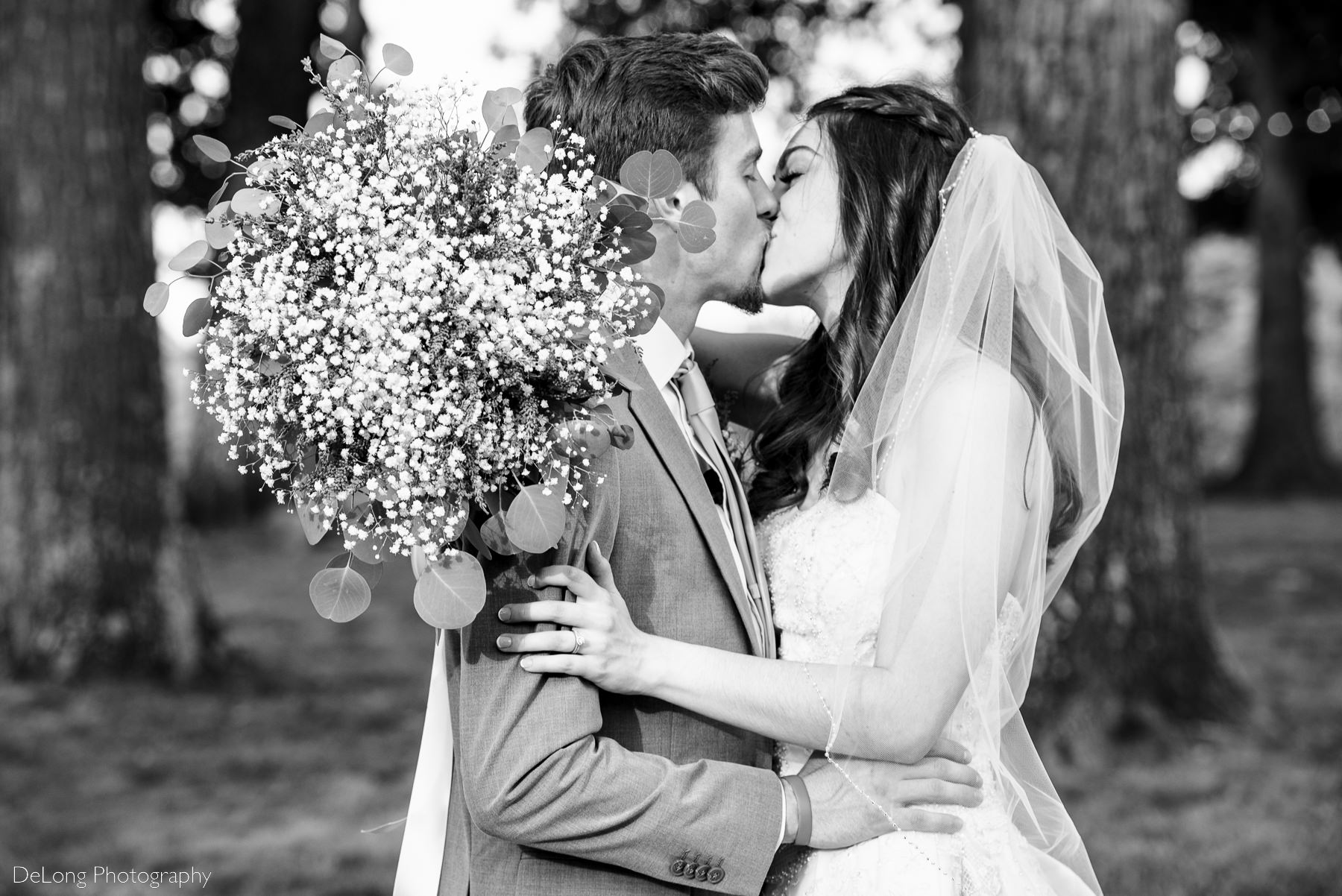 Black and white image of bride and groom kissing near a treeline with her bouquet on the groom's shoulder at Lady Bird Farms by Charlotte Wedding Photographers DeLong Photography
