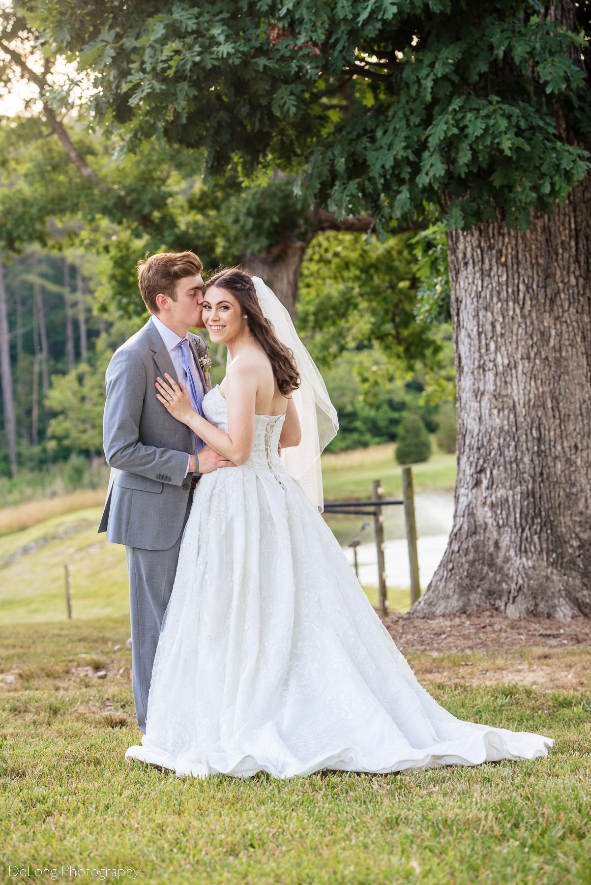 Bride looking back toward the camera smiling while groom kisses her temple  outdoors at Lady Bird Farms by Charlotte Wedding Photographers DeLong Photography